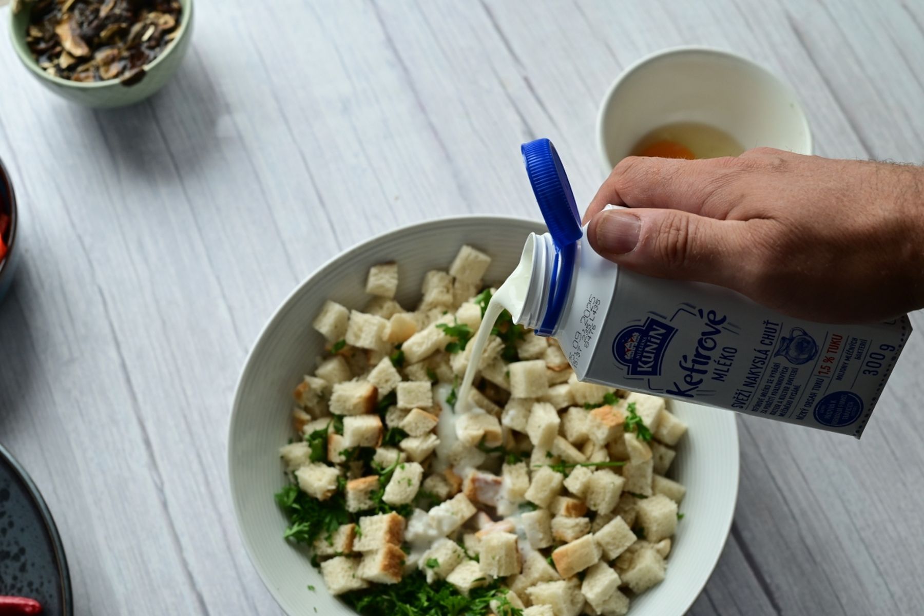 Ruka lijící smetanu z krabice do misky s toustovým chlebem a bylinkami na světlém dřevěném stole, vedle mísa s dalšími surovinami. Recept na kefírový knedlík z hypermarketu Globus od Vojty Poštulky.