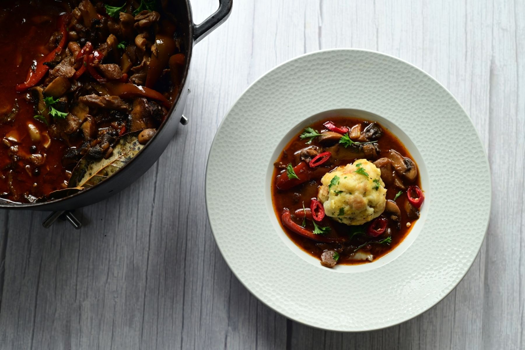 Miska houbového guláše se zeleninou a kefírovým knedlíkem, ozdobená bylinkami a plátky červené chilli papričky, vedle hrnec s gulášem na světlém dřevěném povrchu, podle receptu z hypermarketu Globus od Vojty Poštulky.