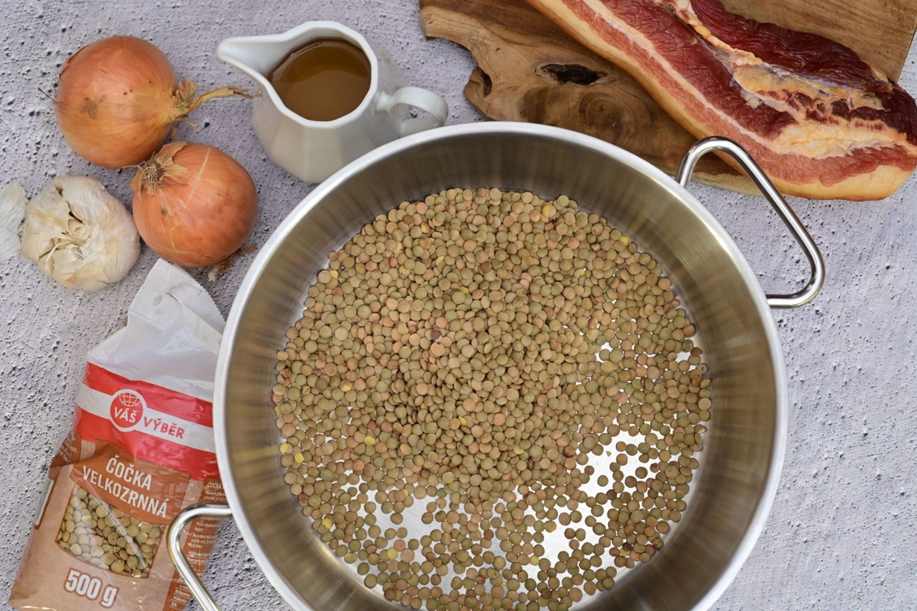 Pohled shora na nerezový hrnec se suchou čočkou, obložený cibulí, česnekem, džbánkem oleje, slaninou a balením čočky o hmotnosti 500 g. Jedná se o recept z nabídky Globus.
