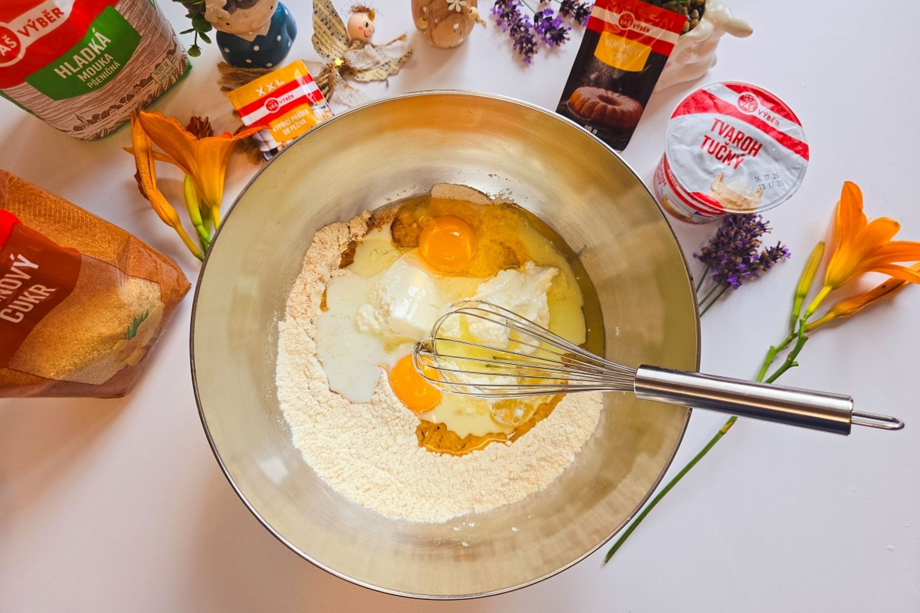 Na bílém povrchu se v misce pomocí metly míchají ingredience na pečení - vejce, mouka a další suroviny, kolem nichž leží mouka, cukr, prášek do pečiva a květiny.