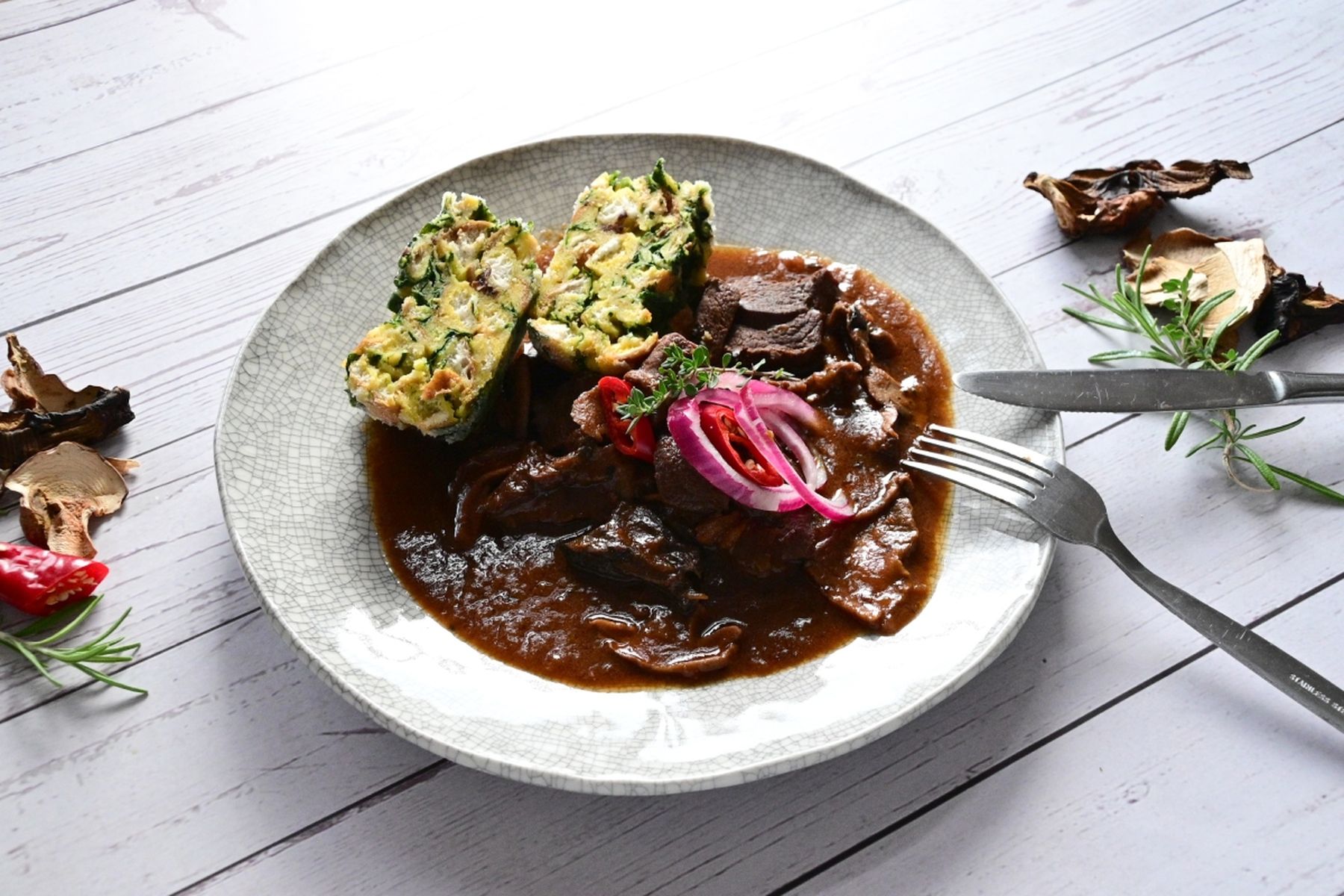 Talíř se srnčím masem v omáčce, ozdobený červenou cibulí a bylinkami, doplněný plátky knedlíku a čerstvým rozmarýnem. Recept připravil Vojta Poštulka.