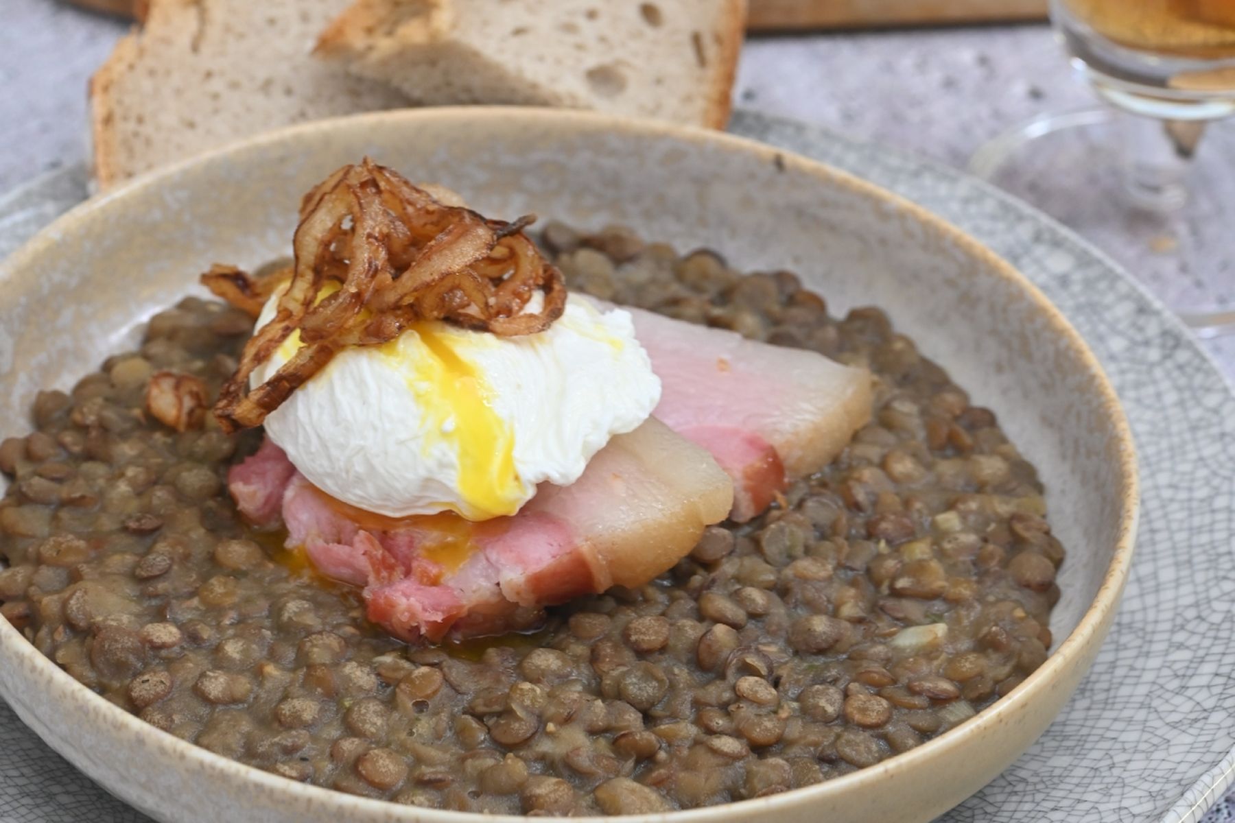 Miska čočky na kyselo podávaná s uzeným bokem, tekutým pošírovaným vejcem a křupavou smaženou cibulkou, v pozadí s pečivem. Jedná se o recept z nabídky Globus.
