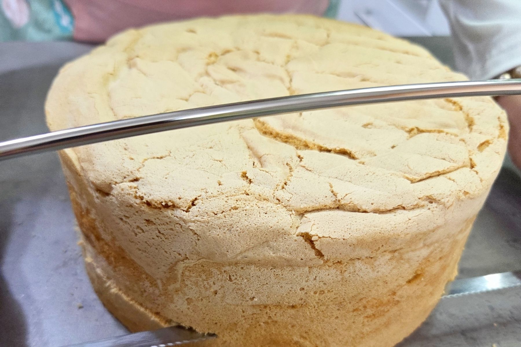 Kulatý, nadýchaný vysoký korpus na kovovém podnosu, na kterém ho někdo pomocí drátěného kráječe na dorty rozřezává vodorovně.