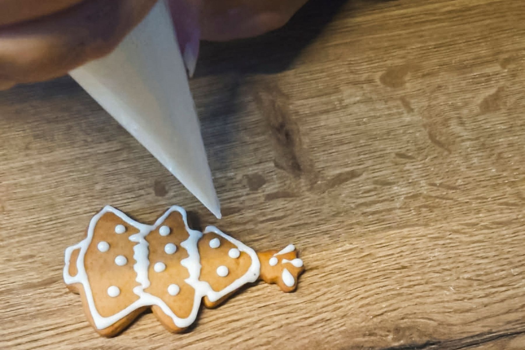 Ruka zdobí perníček ve tvaru vánočního stromečku bílou polevou pomocí zdobicího sáčku na dřevěném povrchu. Recept připravily Ségry v kuchyni.
