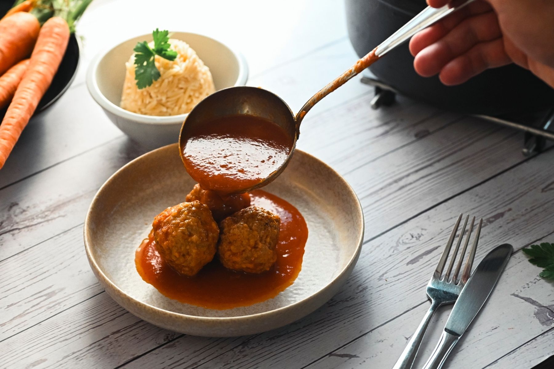 Masové kuličky v rajčatové omáčce na talíři, osoba přelévá další omáčku, v pozadí miska s těstovinovou rýží ozdobenou petrželkou. Recept připravil Vojta Poštulka.