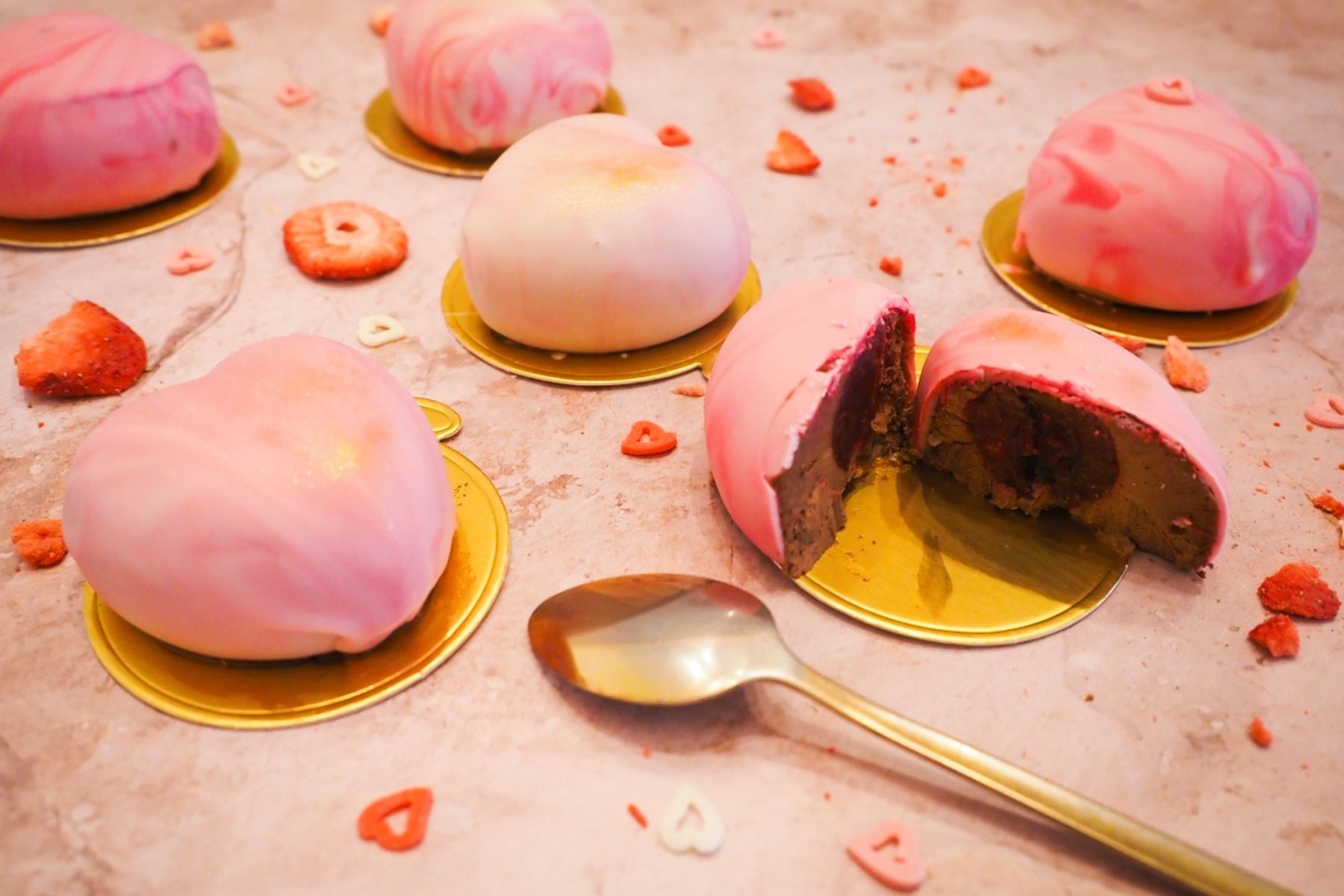 Růžové dezerty ve tvaru srdce na zlatých podložkách s rozsypanými sušenými jahodami a lžičkou na mramorovém povrchu. Jeden je rozkrojený.