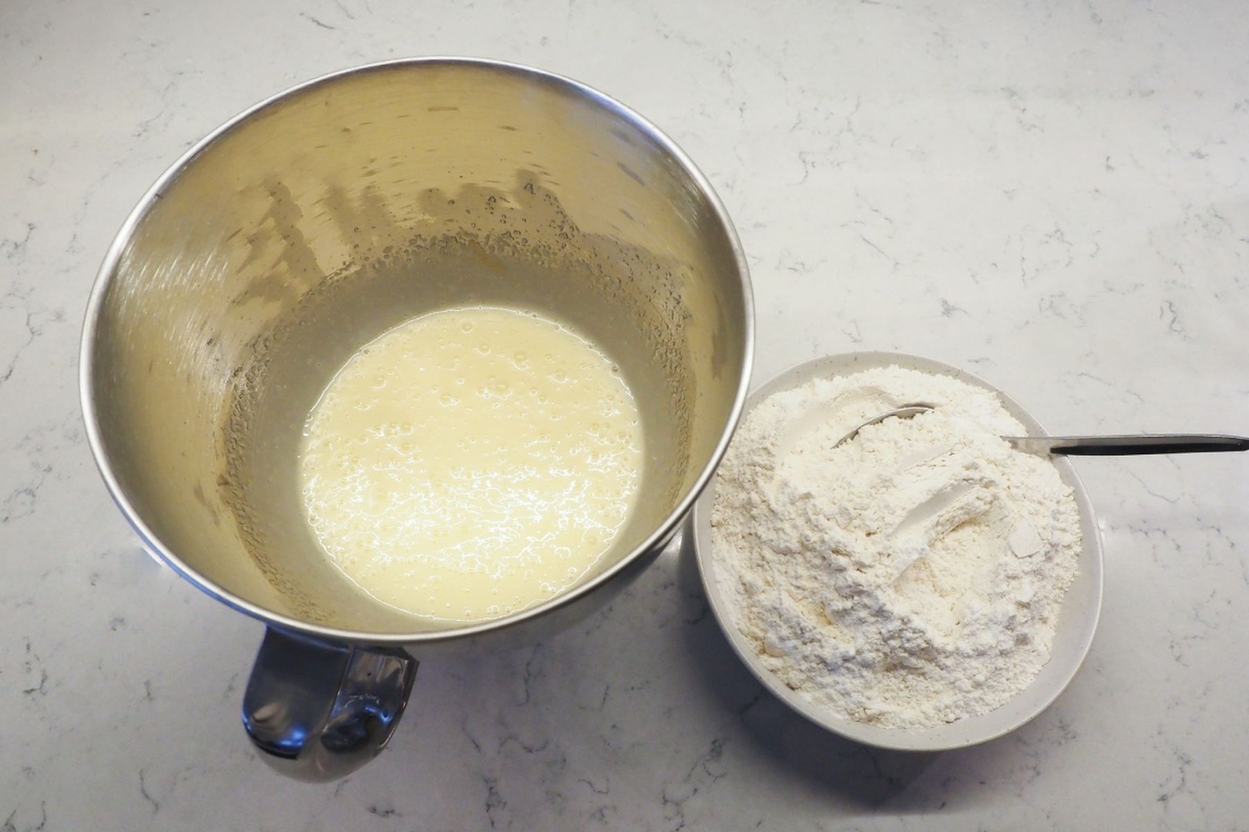 Kovová mísa s vyšlehaným pěnovým těstem vedle bílé misky s moukou a lžící na světlé mramorové pracovní desce. Recept na řezy Dunajské vlny je od Kitchen Affair.