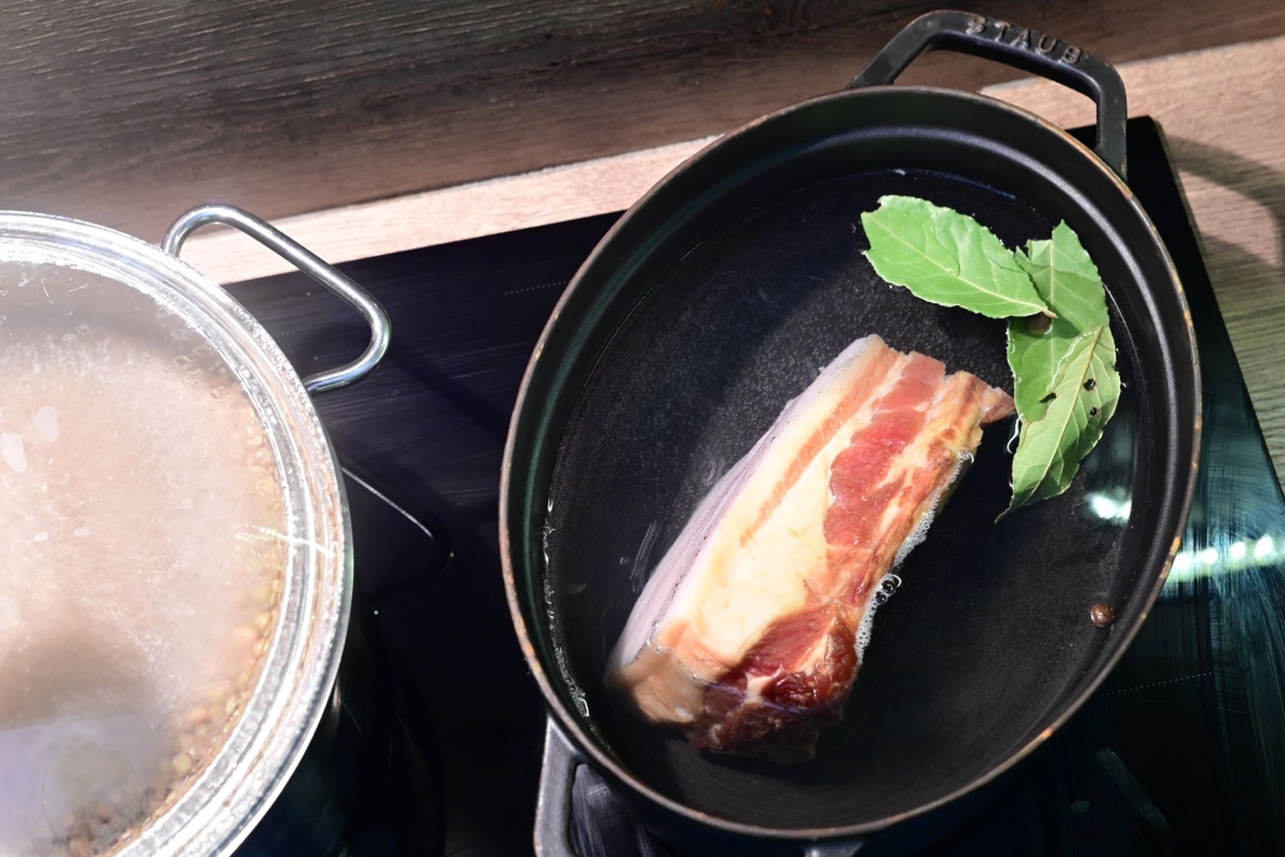 Plát vepřového bůčku a bobkový list provařované v černé litinové pánvi na varné desce, vedle níž probublává zakrytý hrnec. Jedná se o recept z nabídky Globus.