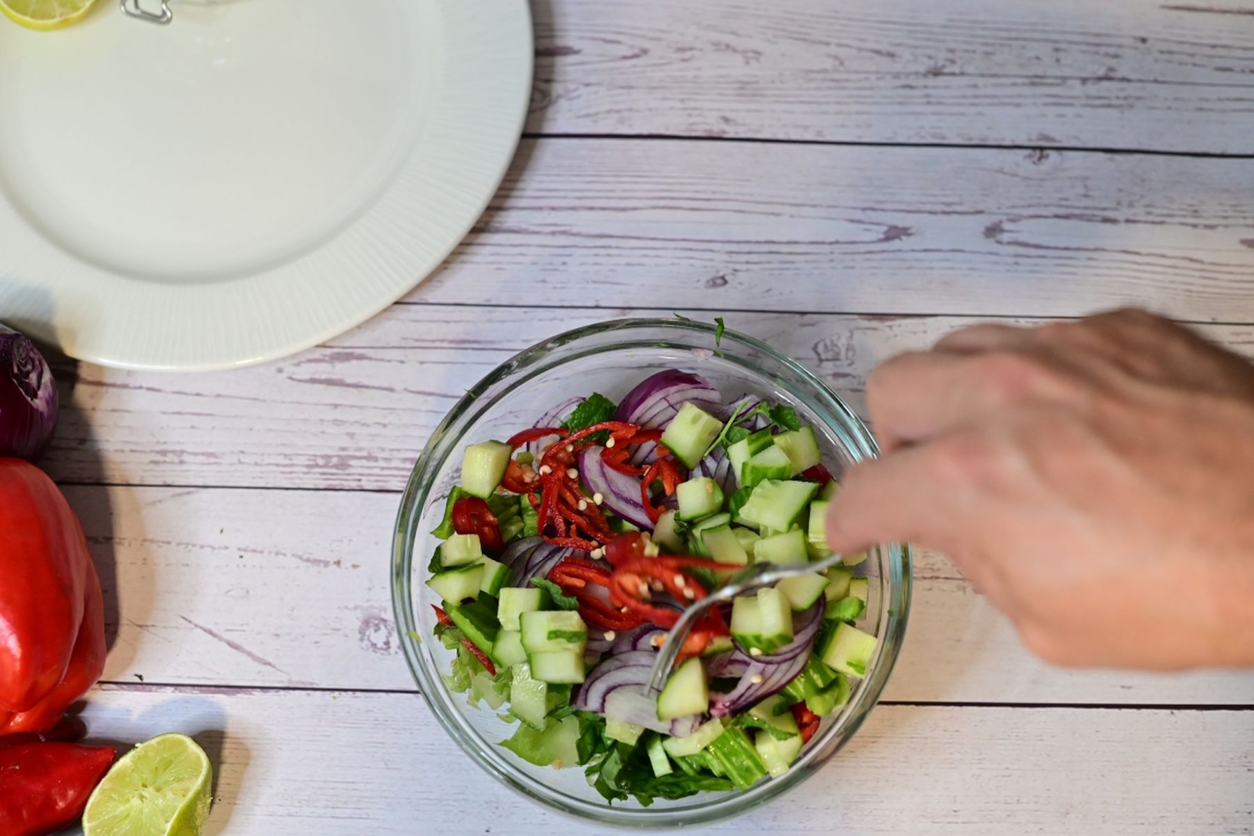 Ruka míchající čerstvý salát z okurek, červené cibule a paprik v skleněné míse na dřevěném stole, vedle talíř a celé kusy zeleniny, součást přípravy receptu na kuřecí kebab z hypermarketu Globus od Vojty Poštulky.