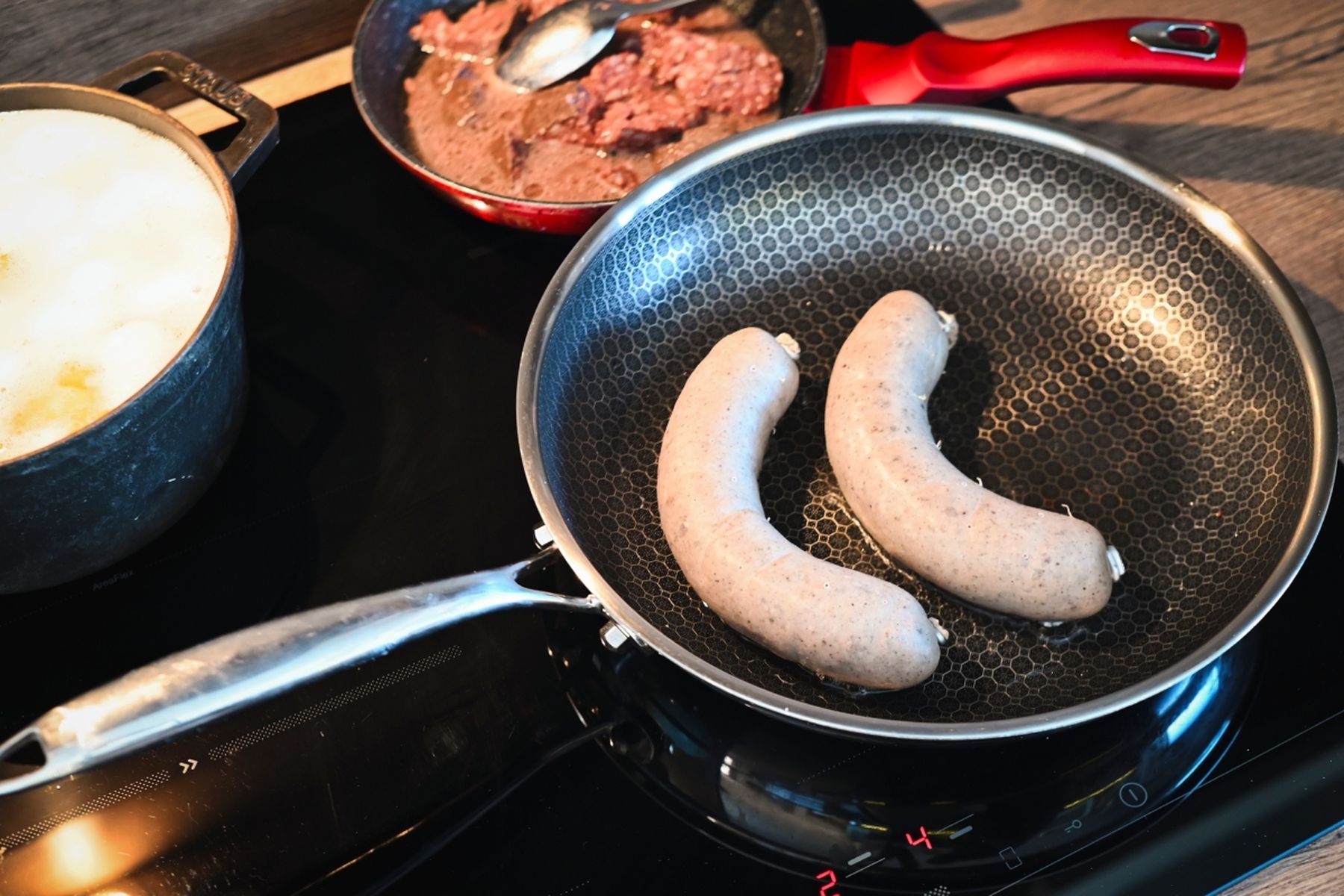 Dvě jitrnice připravované na pánvi na varné desce, v pozadí hrnec a malá pánev s červenou rukojetí a prejtem uvnitř.