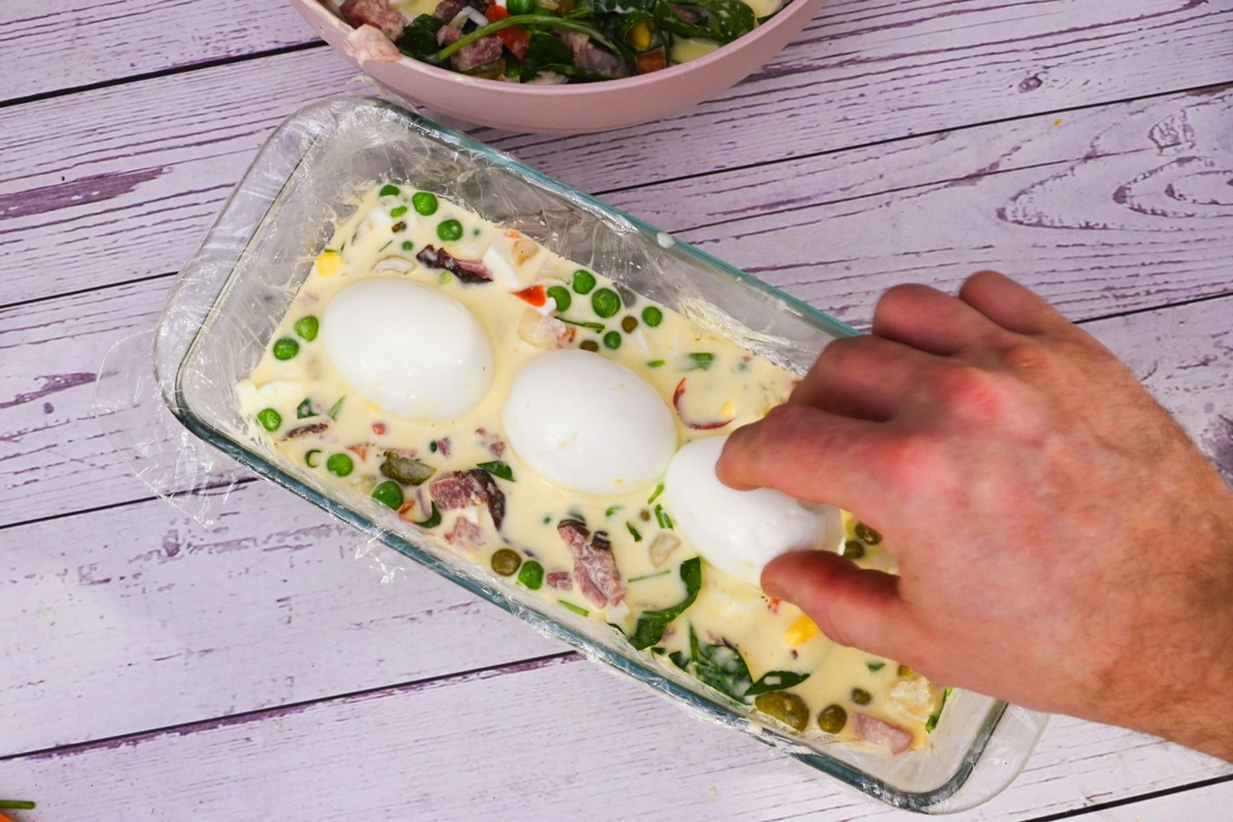 Ruka vkládající vejce do směsi se zeleninou ve formě na chlebíček na světlém dřevěném povrchu, v pozadí miska s ingrediencemi.