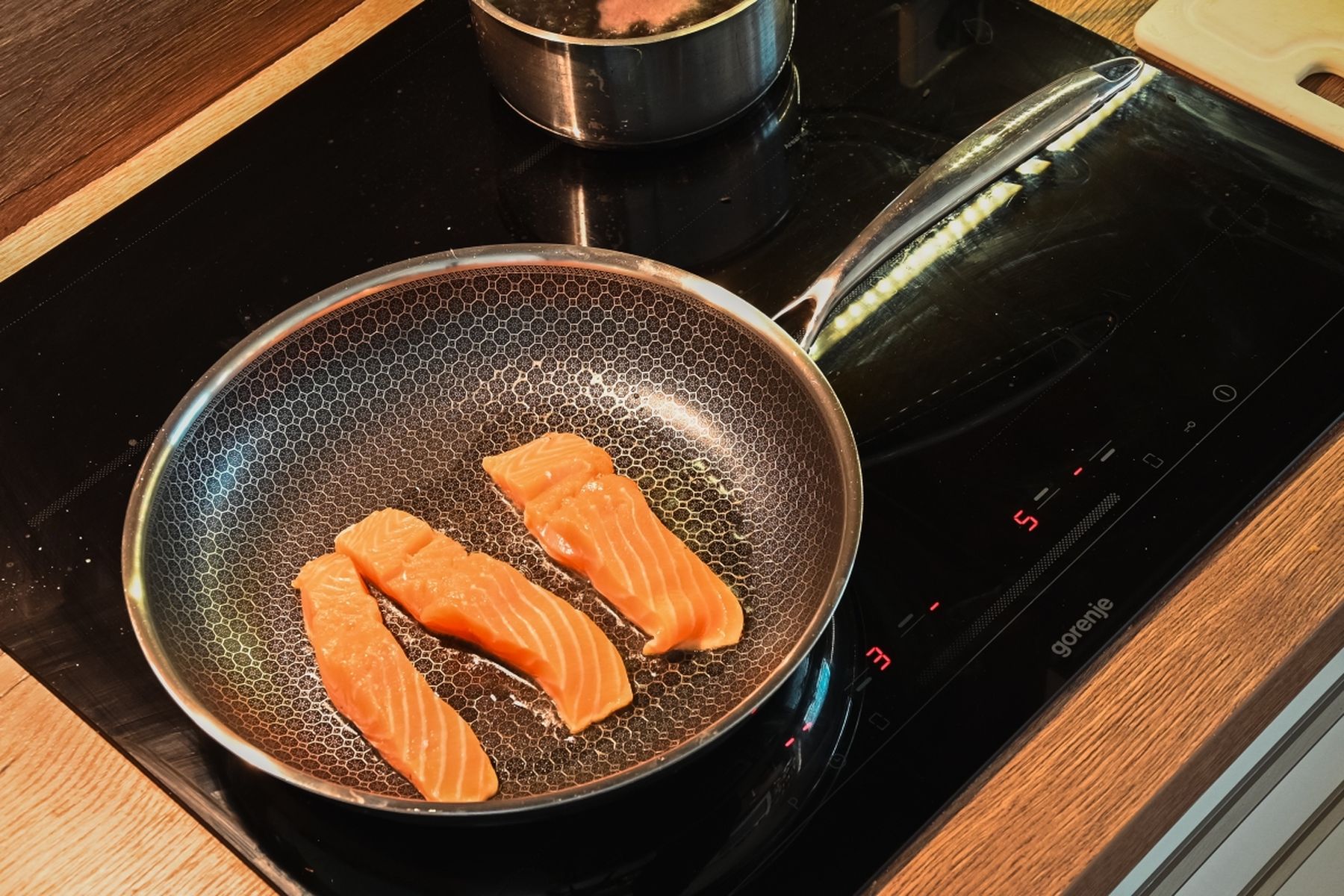 Tři syrové filety z lososa na pánvi na indukční varné desce, v pozadí hrnec. Recept připravil Vojta Poštulka.