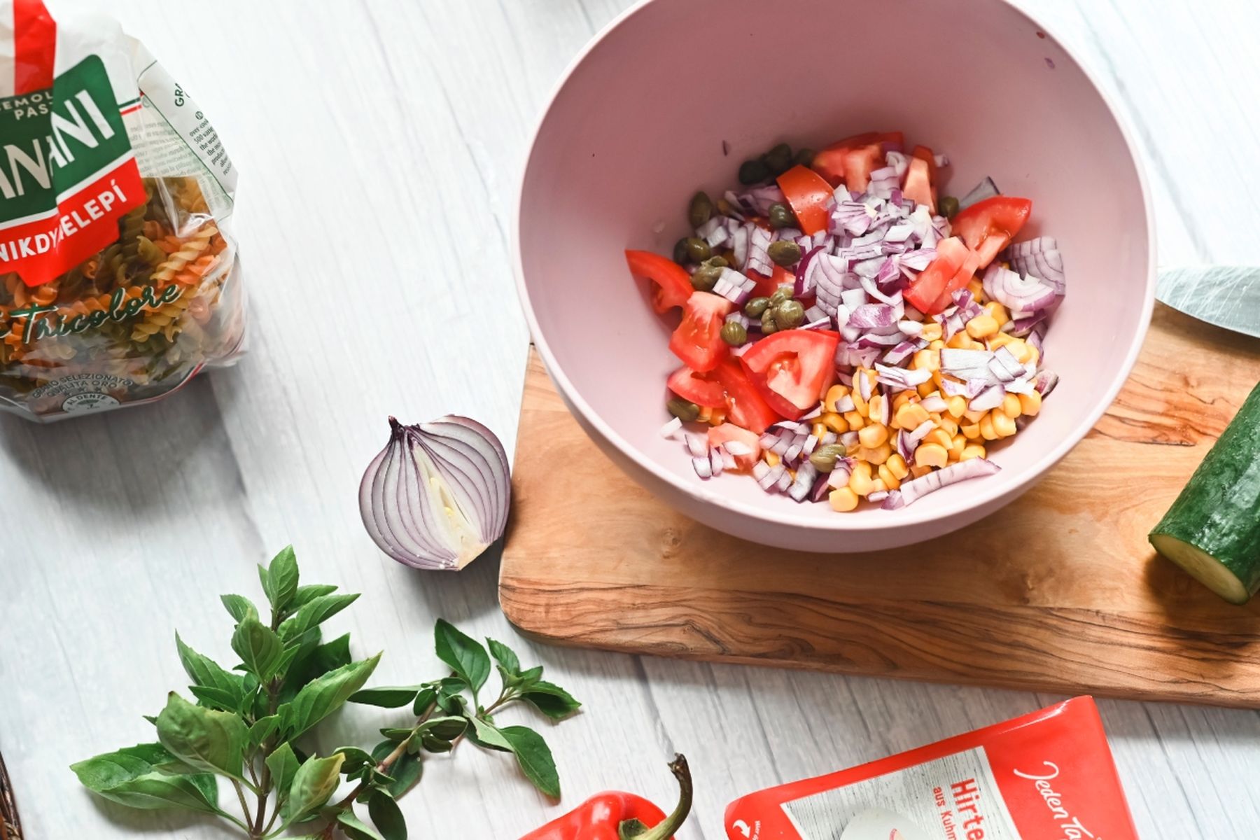 Růžová mísa s nakrájenými rajčaty, červenou cibulí, kapary a kukuřicí na stole, vedle těstoviny, bazalka, paprika a okurka. Recept připravil Vojta Poštulka.