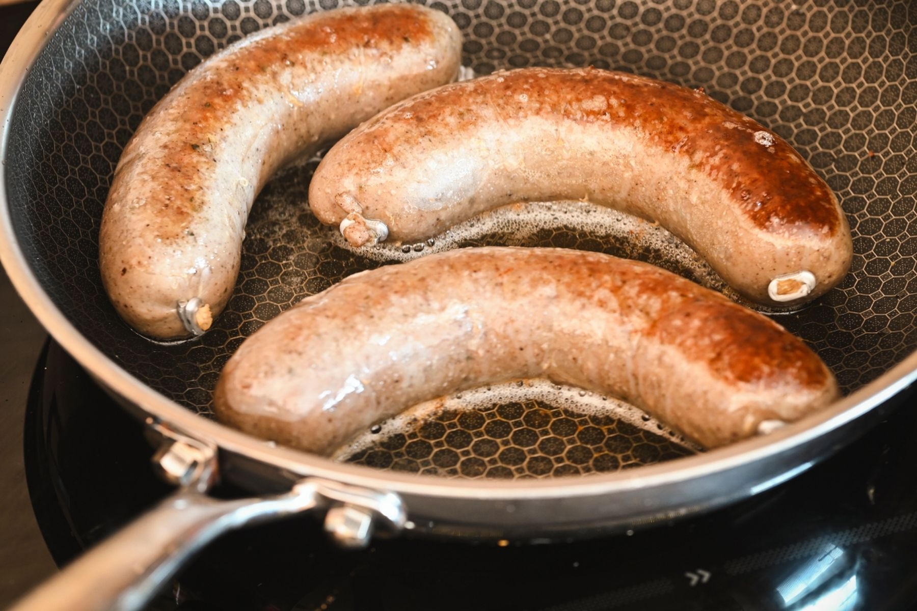 Tři jitrnice opékající se na pánvi, částečně opečené s viditelnými grilovacími stopami, na sporáku.