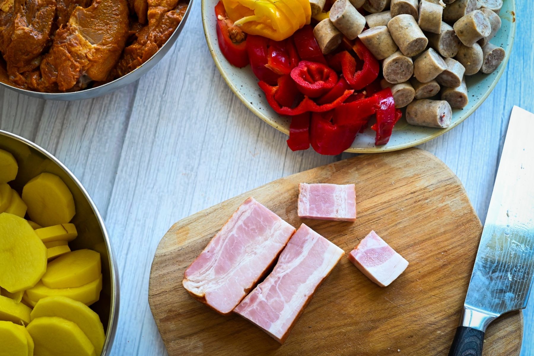 Nakrájená slanina na prkénku s nožem, kolem brambory, naložené maso, klobásy a papriky na světlém dřevěném povrchu. Recept připravil Vojta Poštulka.