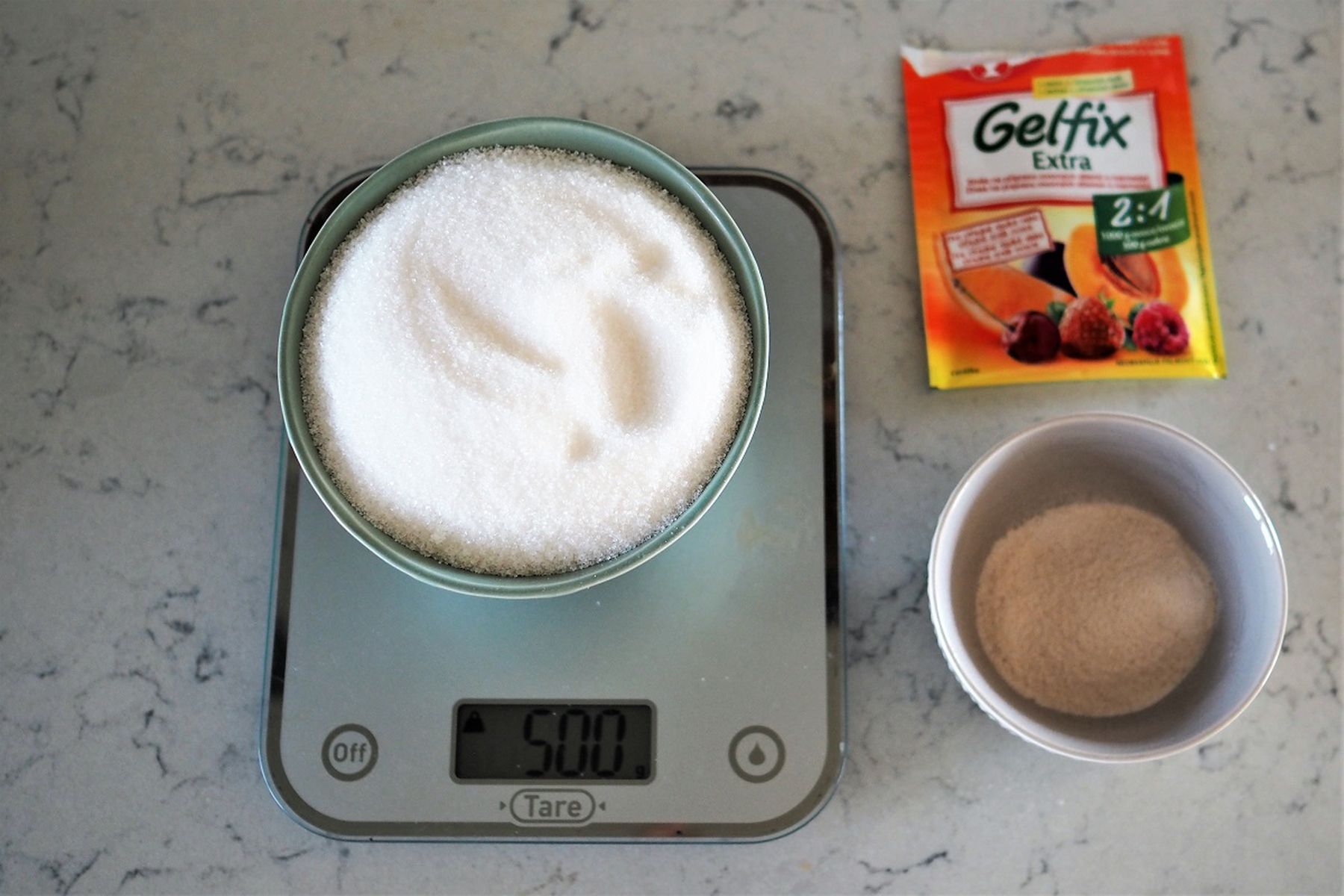 Kuchyňská váha s hodnotou 500 g, na mramorové desce stojí mísa s cukrem, balíček přípravku Gelfix Extra a mísa s práškem.