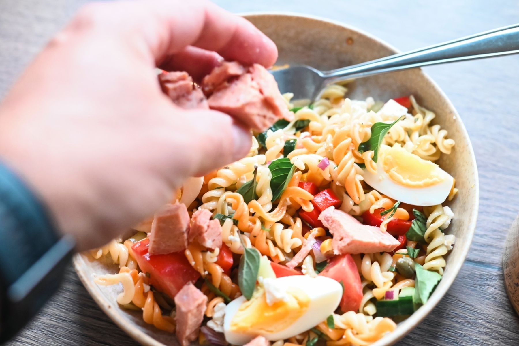 Ruka přidává maso z konzervy z Řeznictví Globus do mísy s těstovinovým salátem z fusilli, vařených vajec, rajčat a bazalky, vedle leží vidlička. Recept připravil Vojta Poštulka.