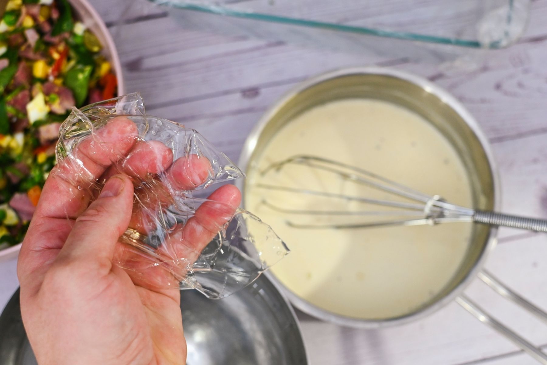Ruka držící plátek želatiny nad mísami na míchání s metličkou a smetanovou směsí, vedle výrazný salát v růžové misce.