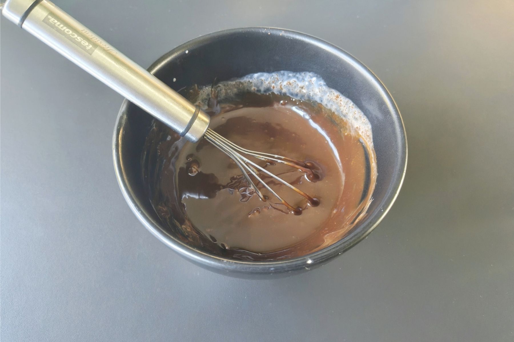 Metla v černé míse s rozpuštěnou čokoládou na hladké tmavé pracovní ploše. Recept připravily Ségry v kuchyni.