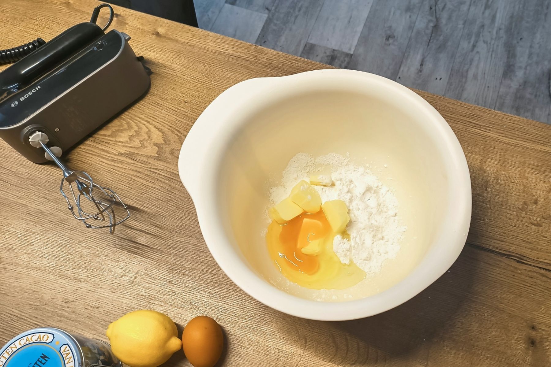 Mísa s moukou, máslem a vejci, vedle citron, vejce a ruční mixér na dřevěné pracovní desce. Recept připravily Ségry v kuchyni.