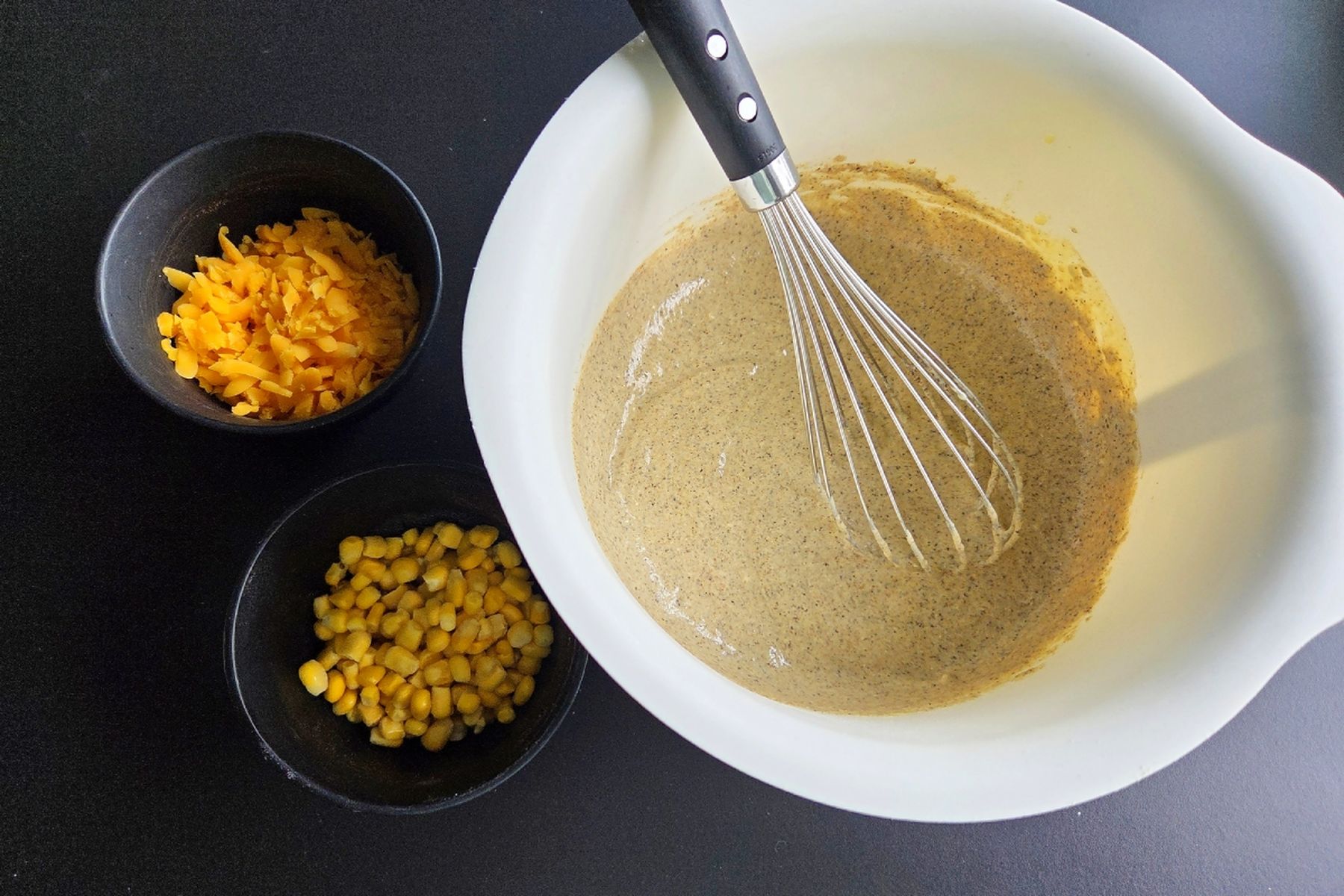 Mísa s těstem a metličkou, vedle malé misky se strouhaným chedarem a kukuřicí na tmavé pracovní desce. Recept připravily Ségry v kuchyni.