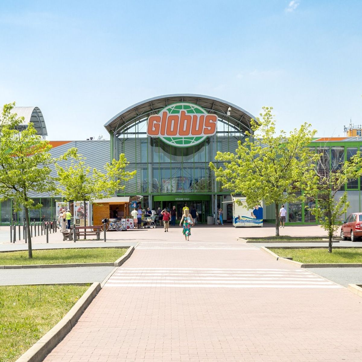 Vchod do supermarketu Globus se skleněnou fasádou, obklopený stromy a zaparkovanými auty; lidé kráčející po chodníku pod jasně modrou oblohou.