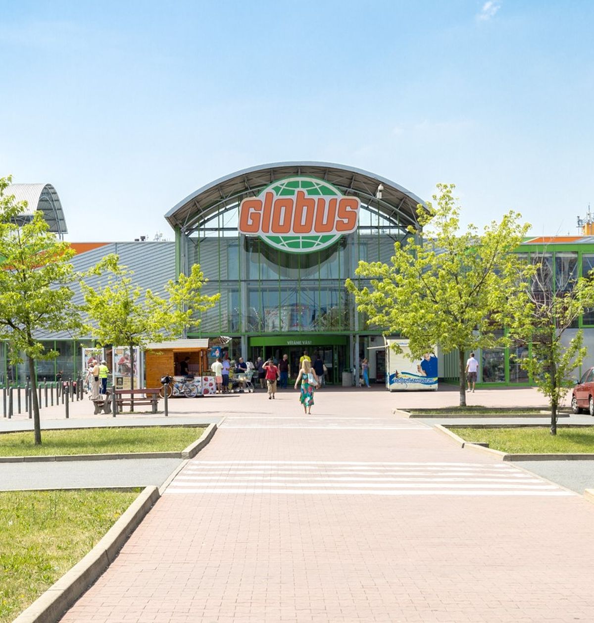 Vchod do supermarketu Globus se skleněnou fasádou, obklopený stromy a zaparkovanými auty; lidé kráčející po chodníku pod jasně modrou oblohou.