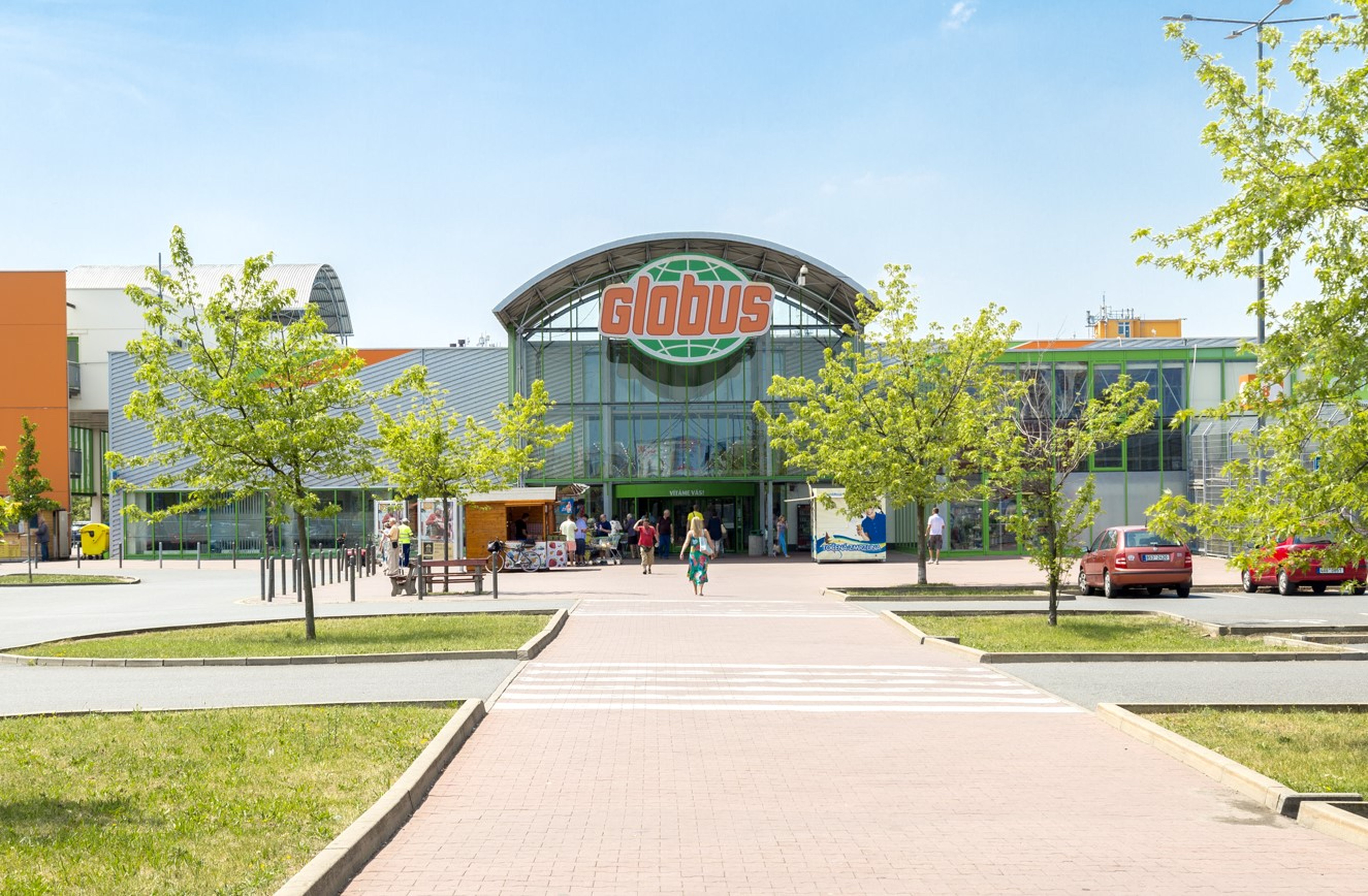 Vchod do supermarketu Globus se skleněnou fasádou, obklopený stromy a zaparkovanými auty; lidé kráčející po chodníku pod jasně modrou oblohou.