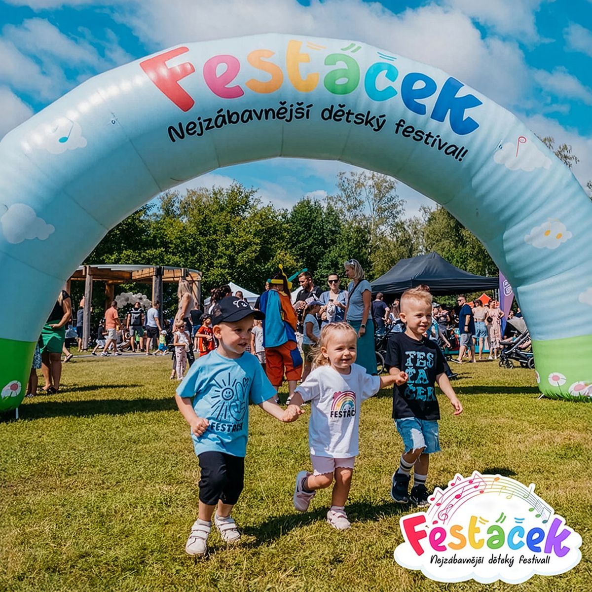Tři děti se drží za ruce a běží pod barevným obloukem na živém venkovním festivalu; v pozadí jsou lidé a panuje slunečný den.