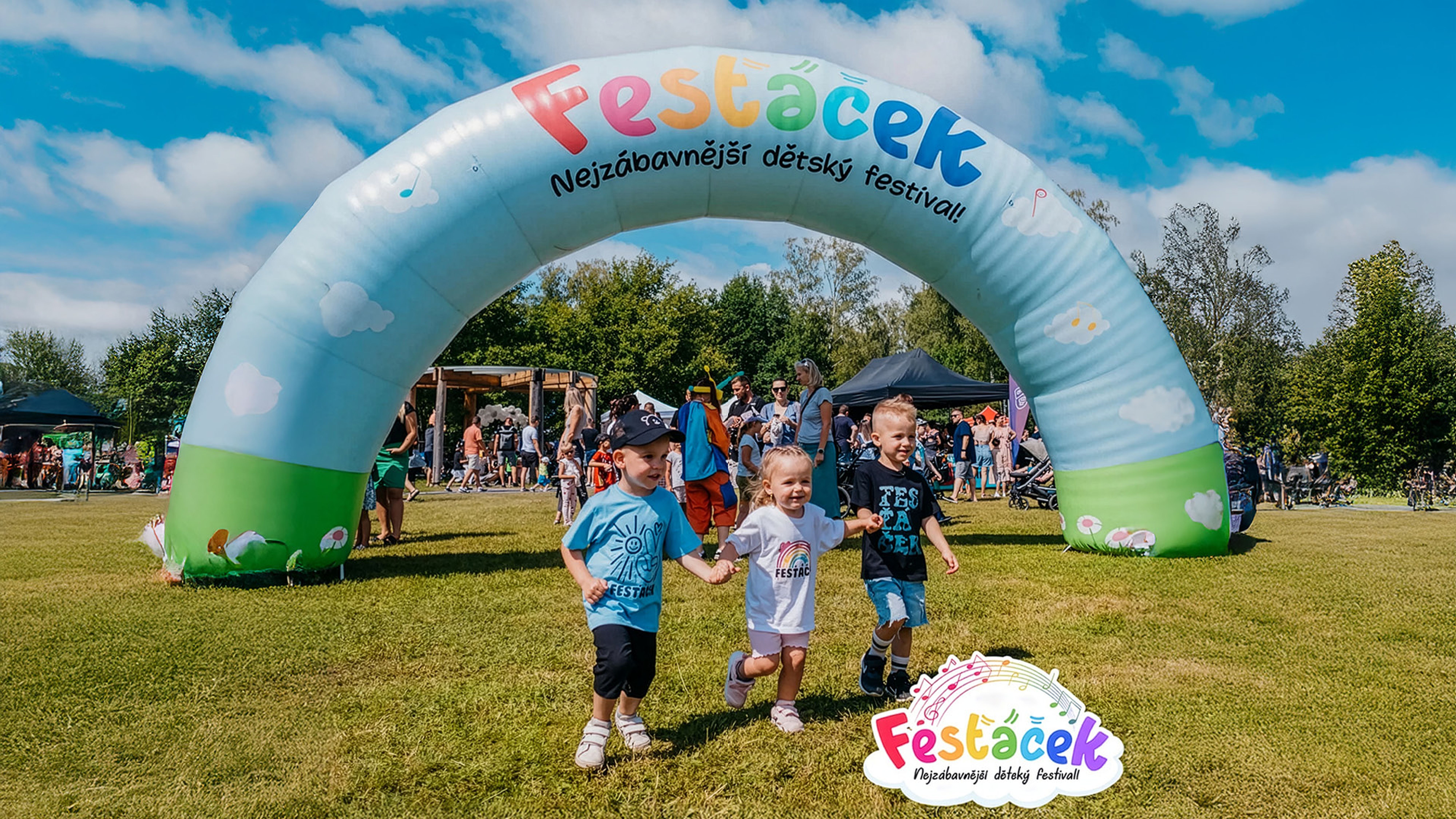 Tři děti se drží za ruce a běží pod barevným obloukem na živém venkovním festivalu; v pozadí jsou lidé a panuje slunečný den.