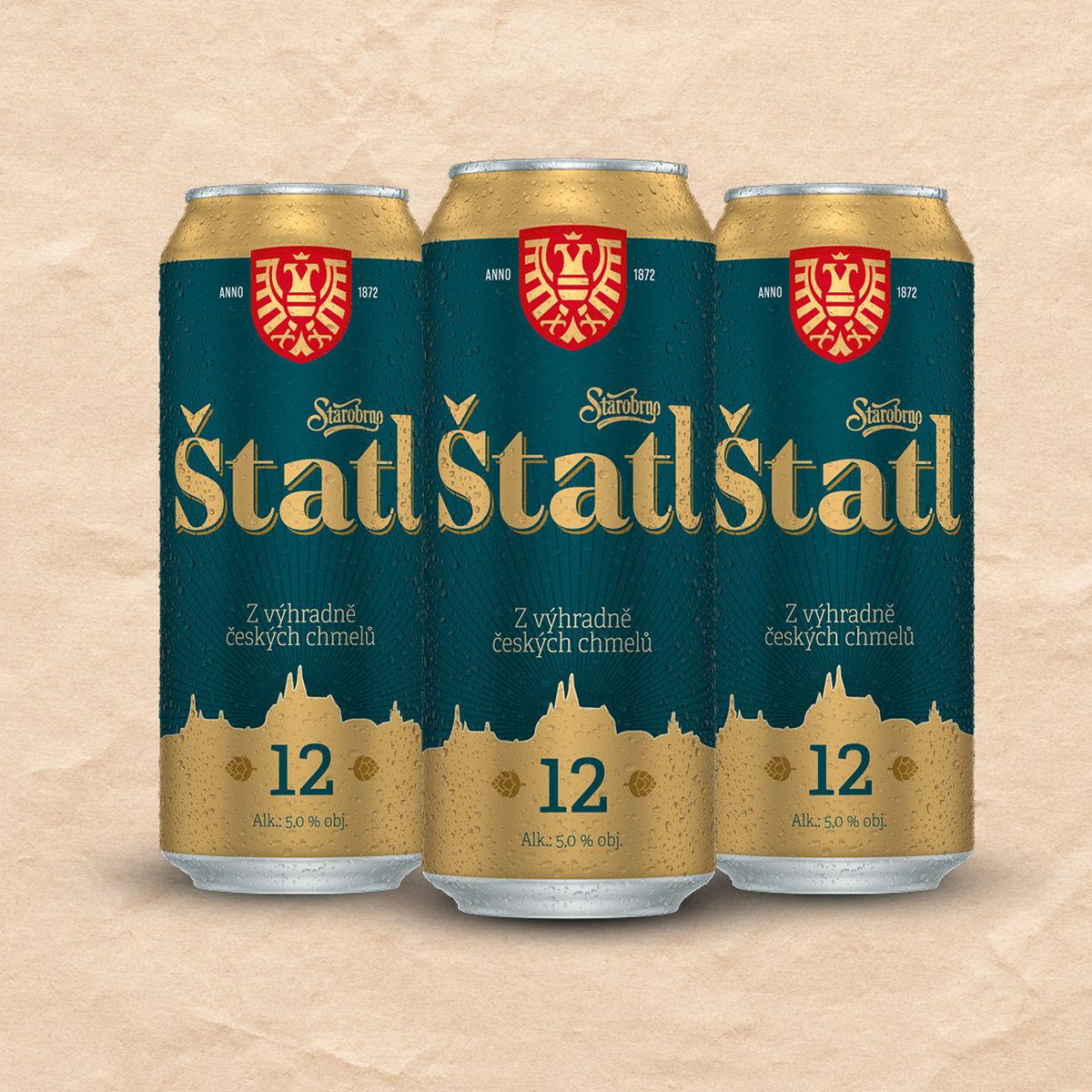 Tři plechovky piva s textem Štatl, tmavě zelenými etiketami a zlatými akcenty na světlém texturovaném pozadí.