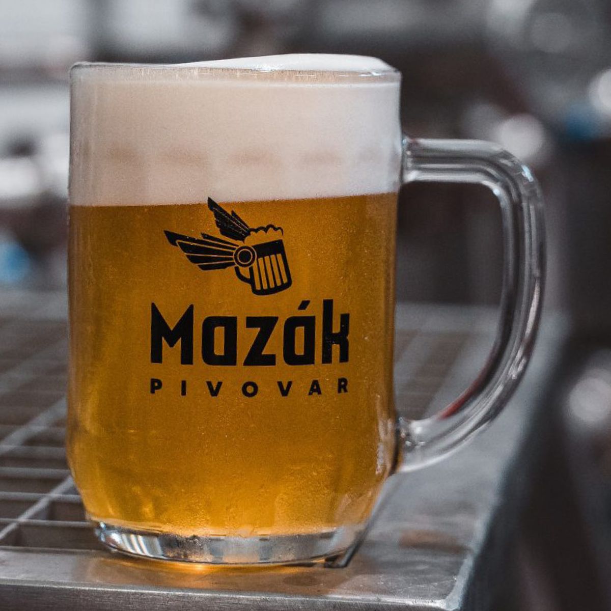 Skleněný půllitr s pivem s bohatou pěnou stojí na kovovém povrchu; na sklenici je logo „Mazák Pivovar“ a pozadí je rozmazané.