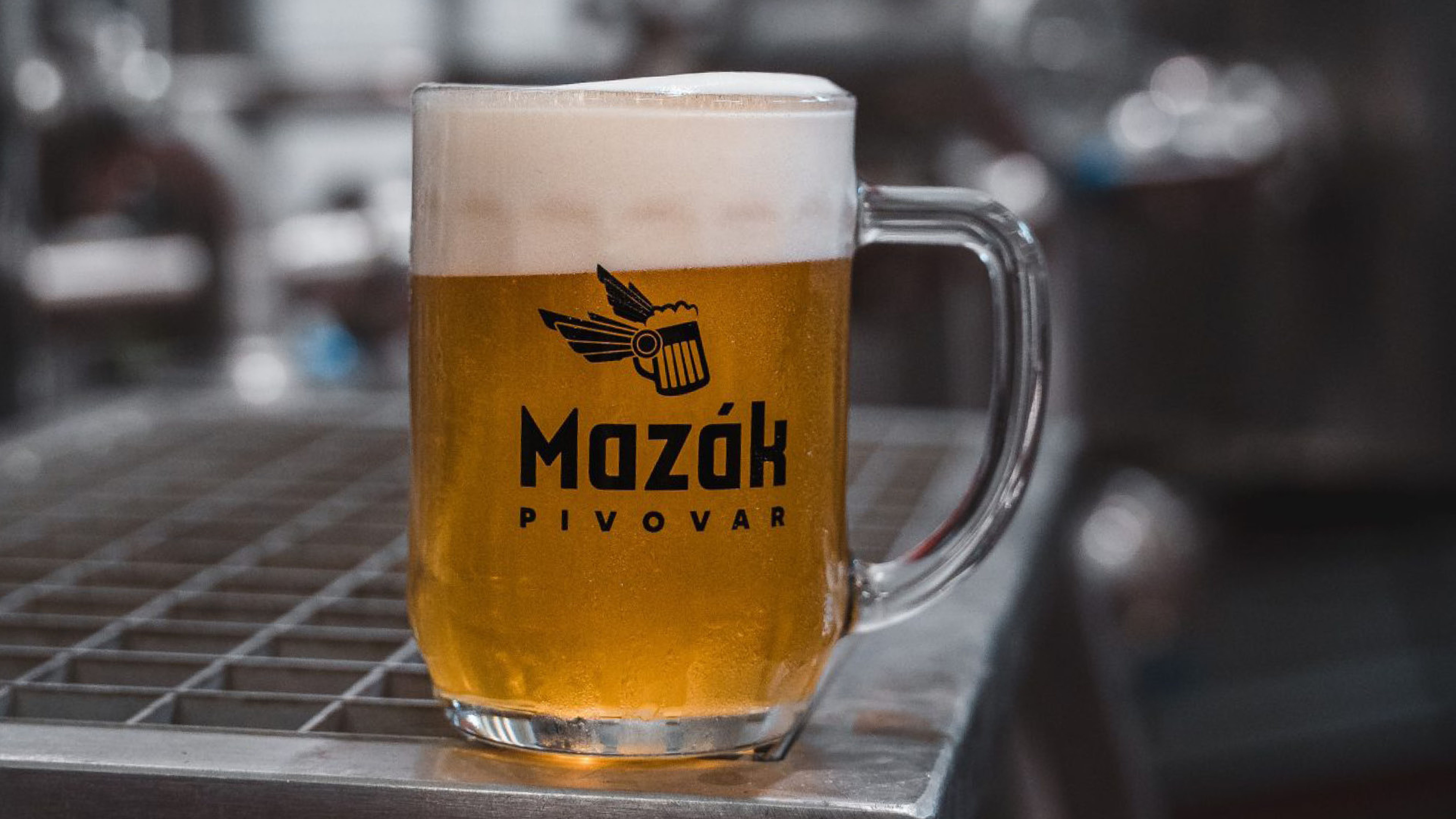 Skleněný půllitr s pivem s bohatou pěnou stojí na kovovém povrchu; na sklenici je logo „Mazák Pivovar“ a pozadí je rozmazané.