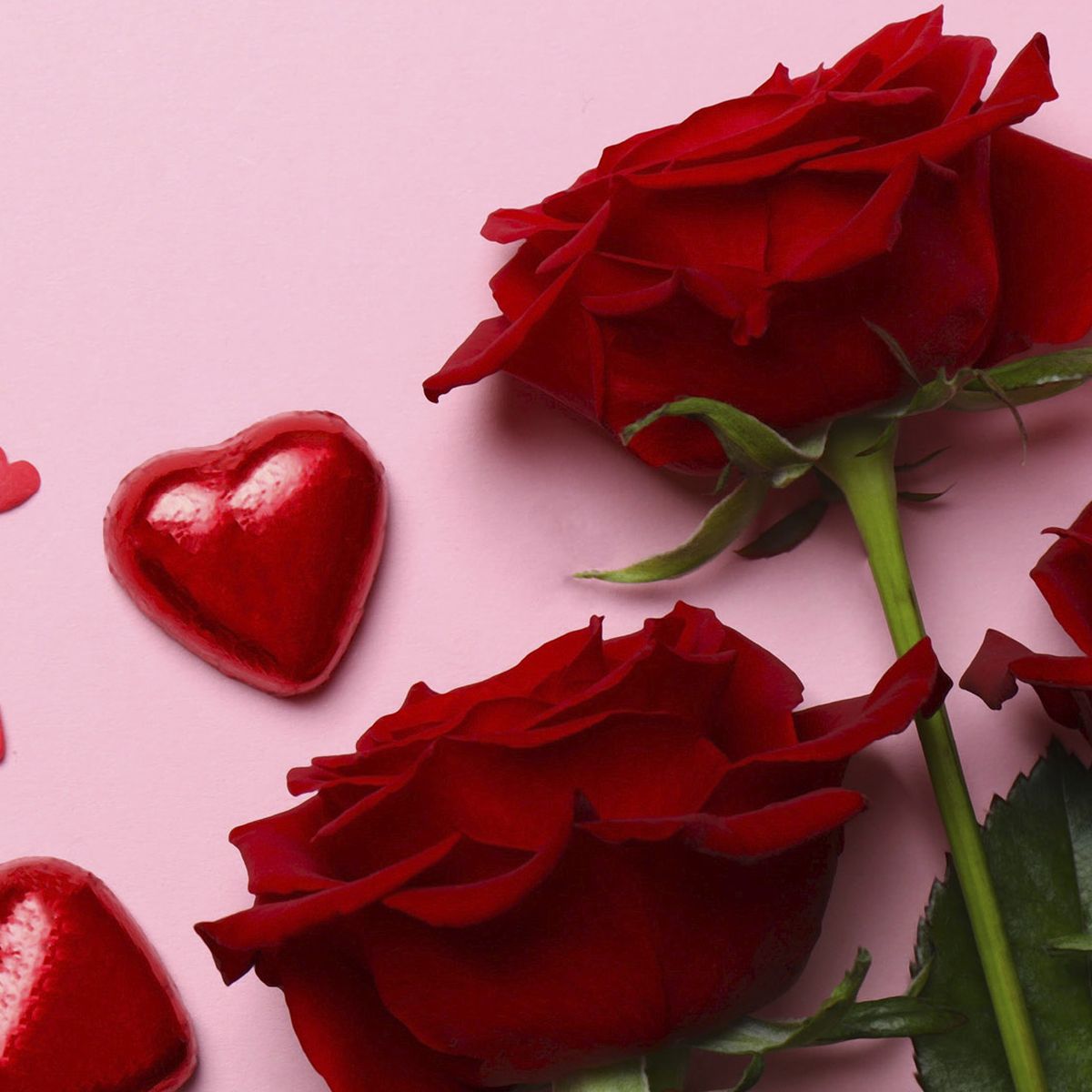 Červené růže se zelenými listy a čokoládami ve tvaru srdce na růžovém pozadí, doplněné malými červenými ozdobami srdce.