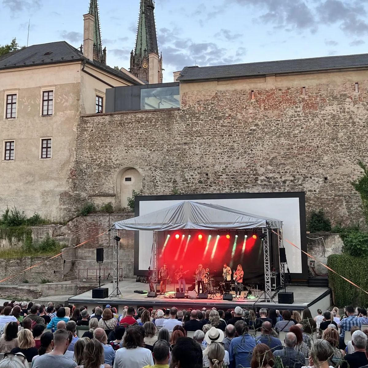 Venkovní koncert s velkým publikem před historickou budovou, na jevišti osvětleném červenými světly proti jasné večerní obloze.