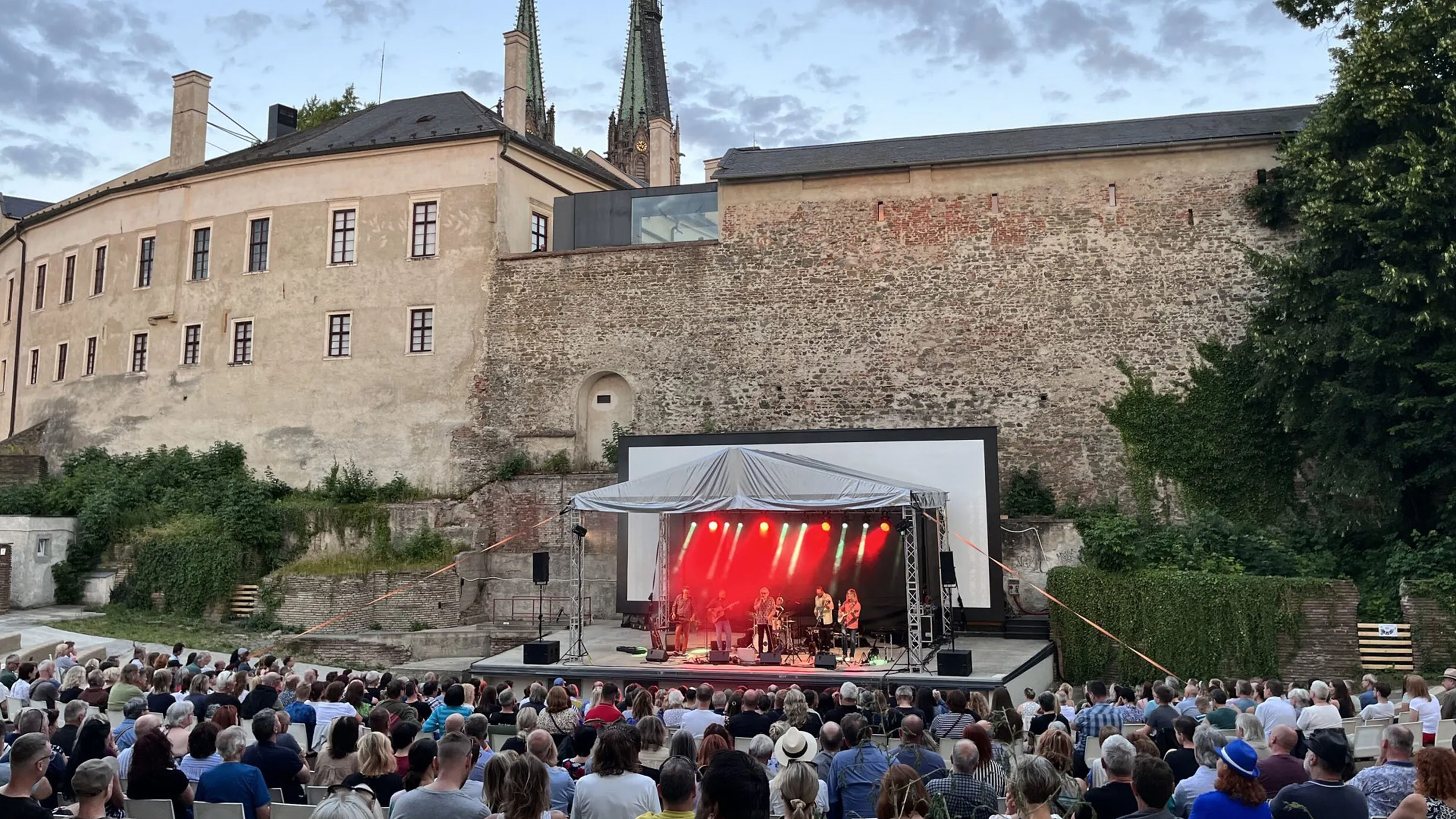 Venkovní koncert s velkým publikem před historickou budovou, na jevišti osvětleném červenými světly proti jasné večerní obloze.
