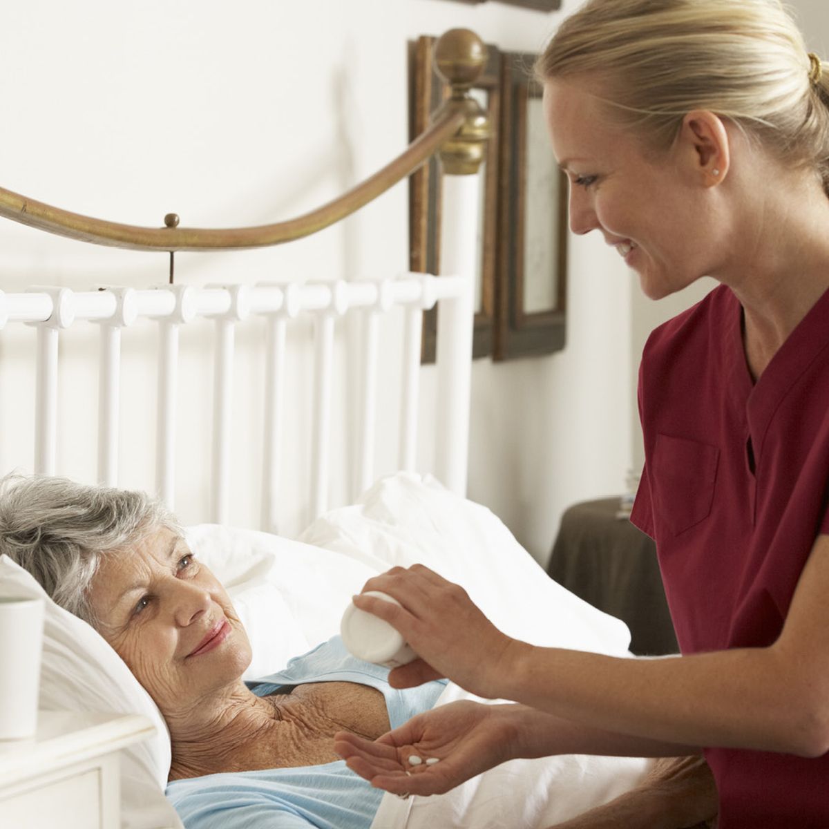 Zdravotní sestra v červeném oděvu podává léky usmívající se starší ženě ležící v posteli v ložnici.