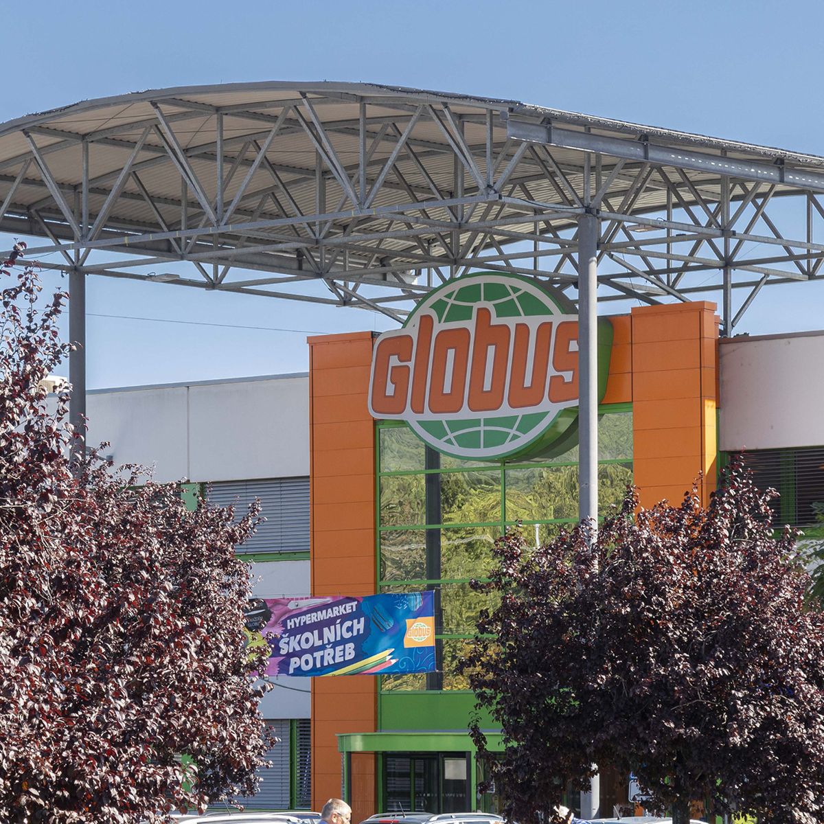 Exteriér hypermarketu Globus s velkým logem, oranžovou a zelenou fasádou a kovovým přístřeškem, obklopený stromy.