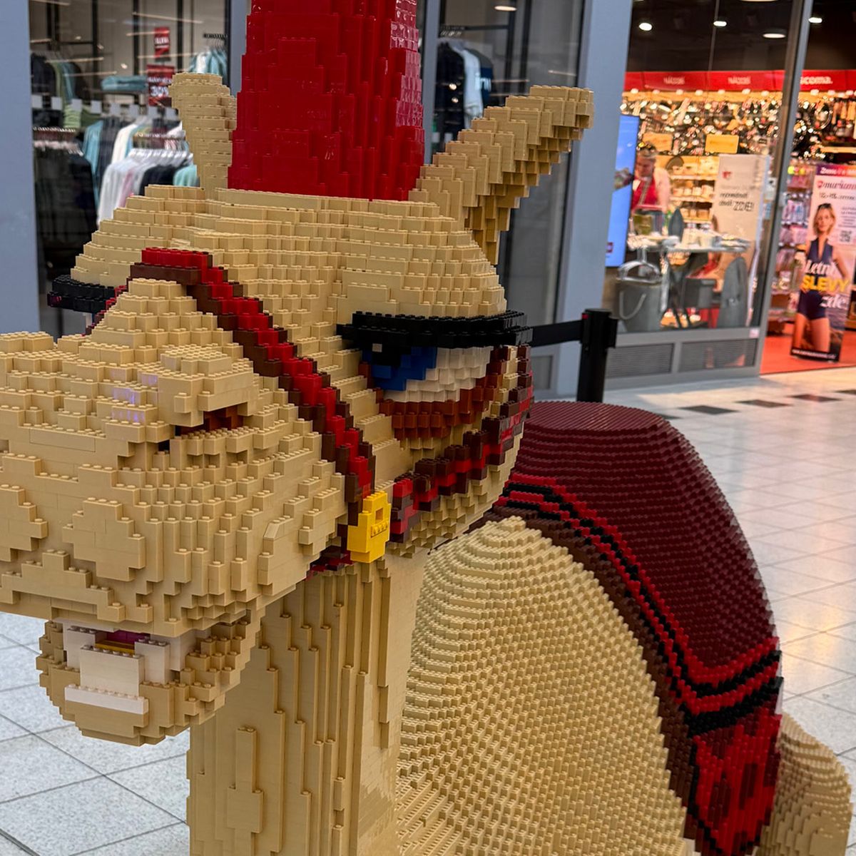 Lego socha velblouda s červeným sedlem a čelenkou vystavená v nákupním středisku.