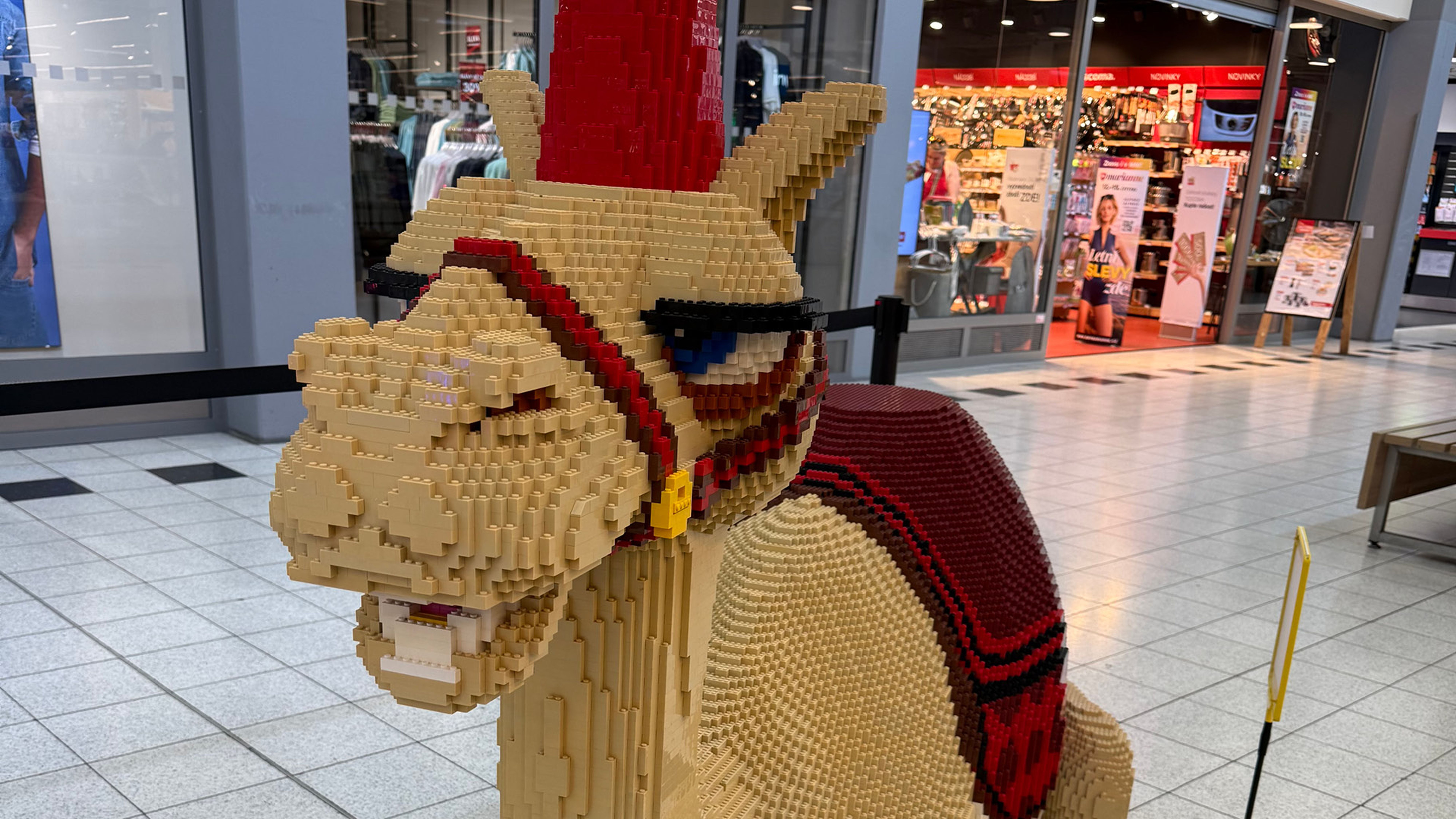 Lego socha velblouda s červeným sedlem a čelenkou vystavená v nákupním středisku.