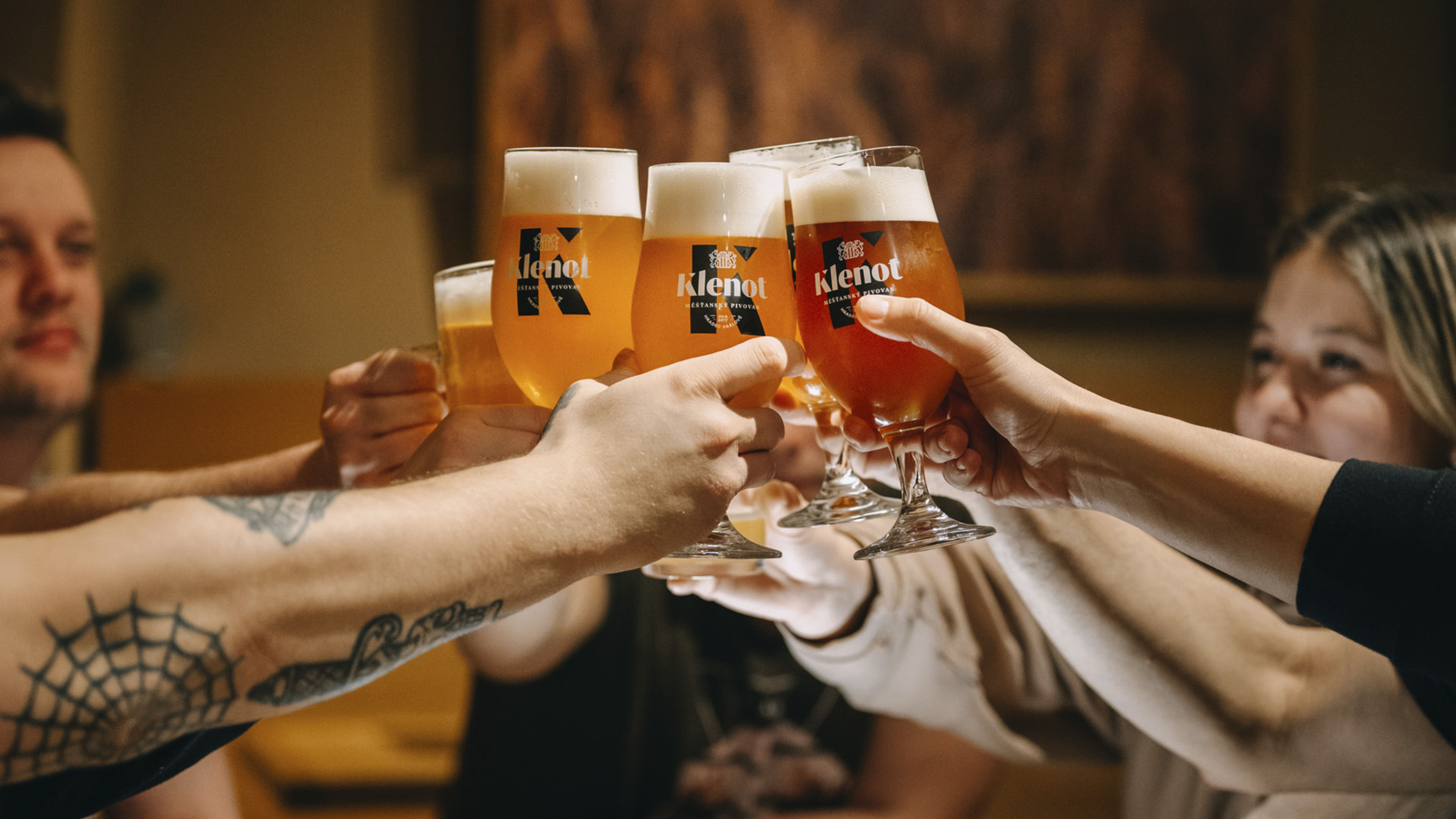 Skupina lidí si připíjí sklenicemi piva s etiketou „Klenot“ v útulném, tlumeně osvětleném prostředí.