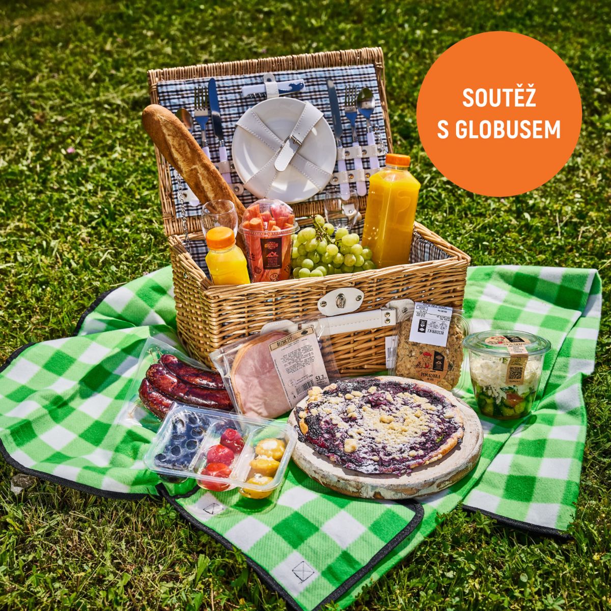 Piknikový koš s bagetou, pomerančovým džusem, hroznovým vínem, nakrájenou zeleninou, klobásami, borůvkovým koláčem, šunkou, šopským salátem a antipasti na zelené kostkované dece na trávě.