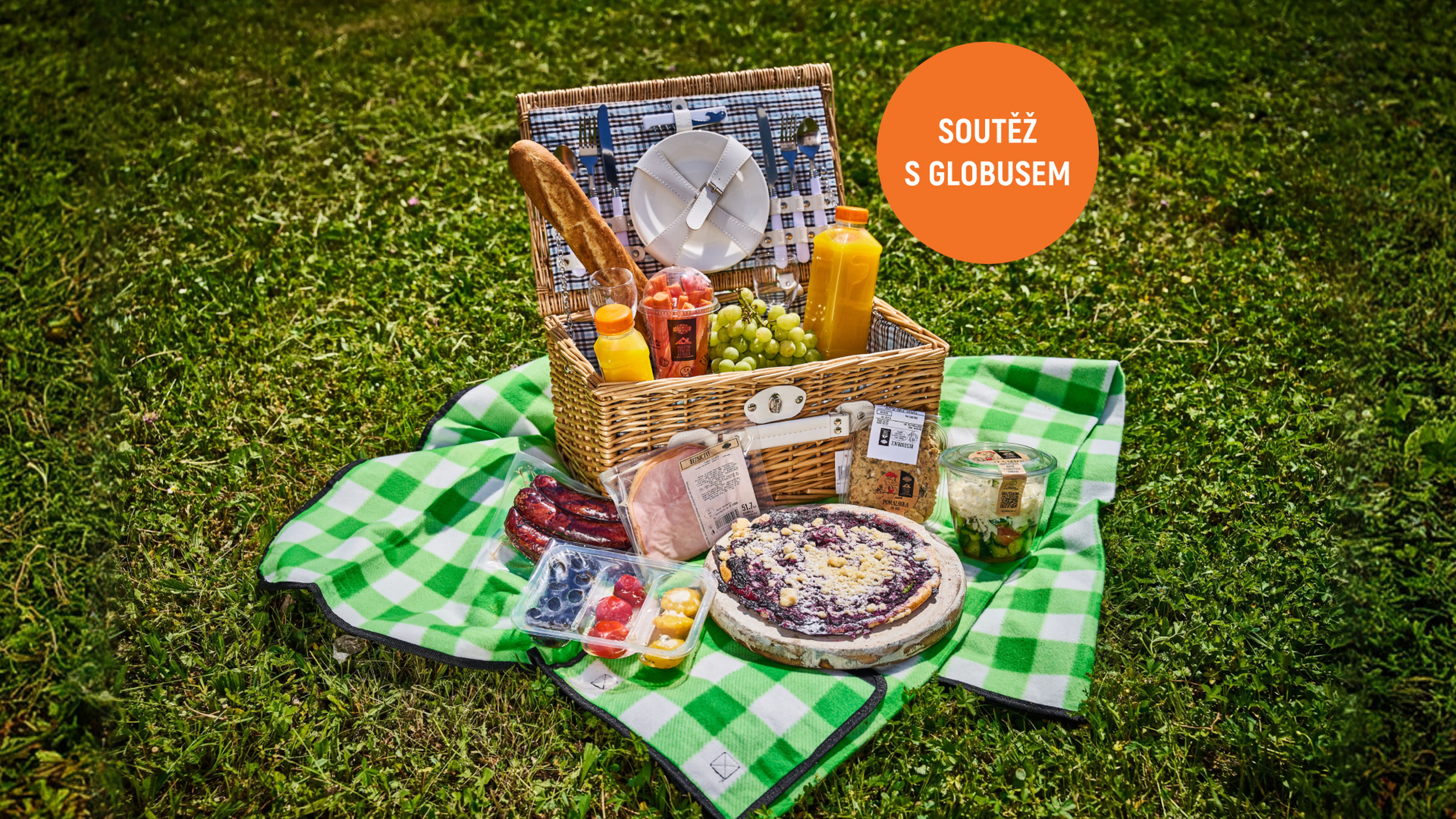 Piknikový koš s bagetou, pomerančovým džusem, hroznovým vínem, nakrájenou zeleninou, klobásami, borůvkovým koláčem, šunkou, šopským salátem a antipasti na zelené kostkované dece na trávě.
