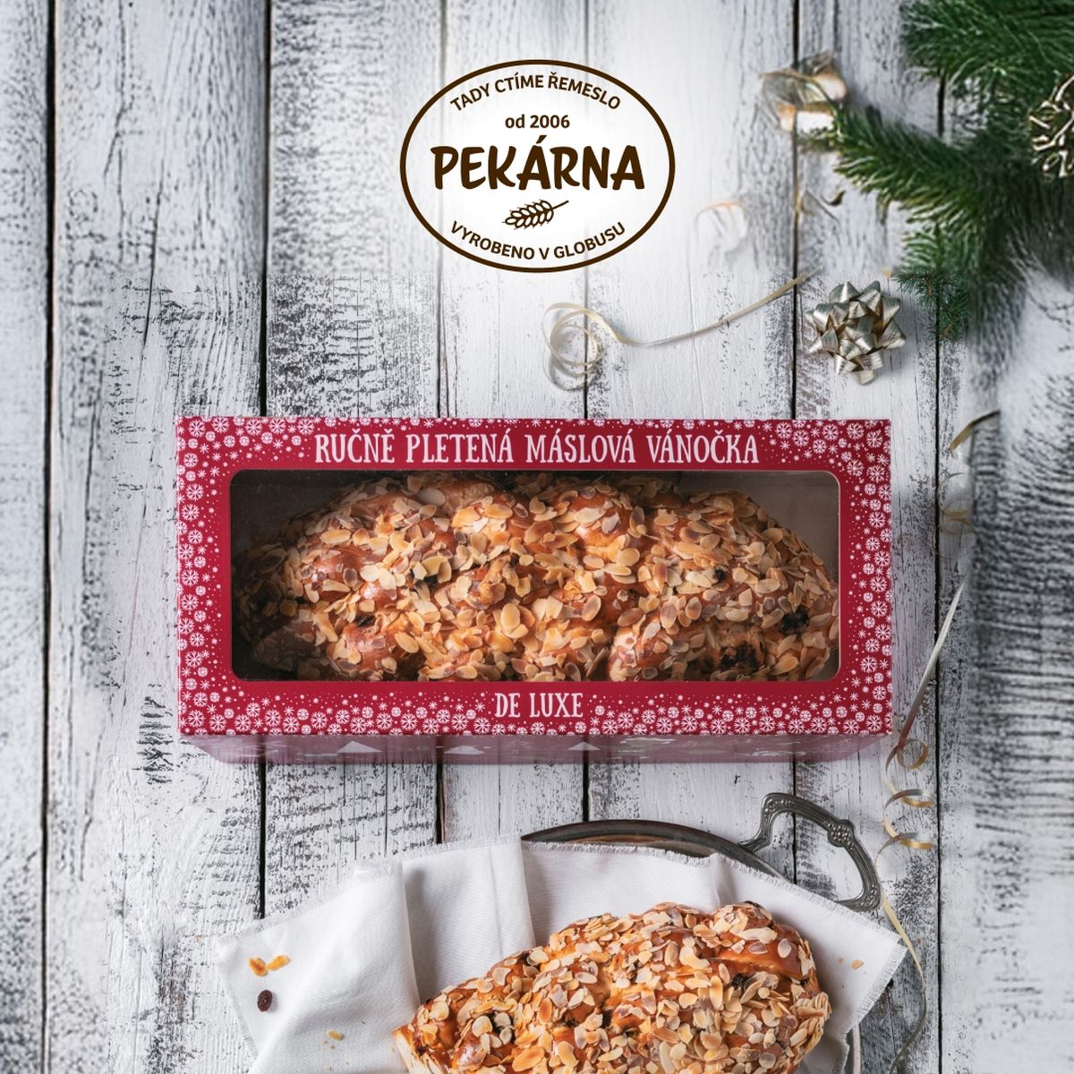 Máslová vánočka de luxe v červené papírové krabici, kus rozkrojené vánočky na bílé látce a tácu, zelené jehličí s ozdobami, vše na dřevěném pozadí