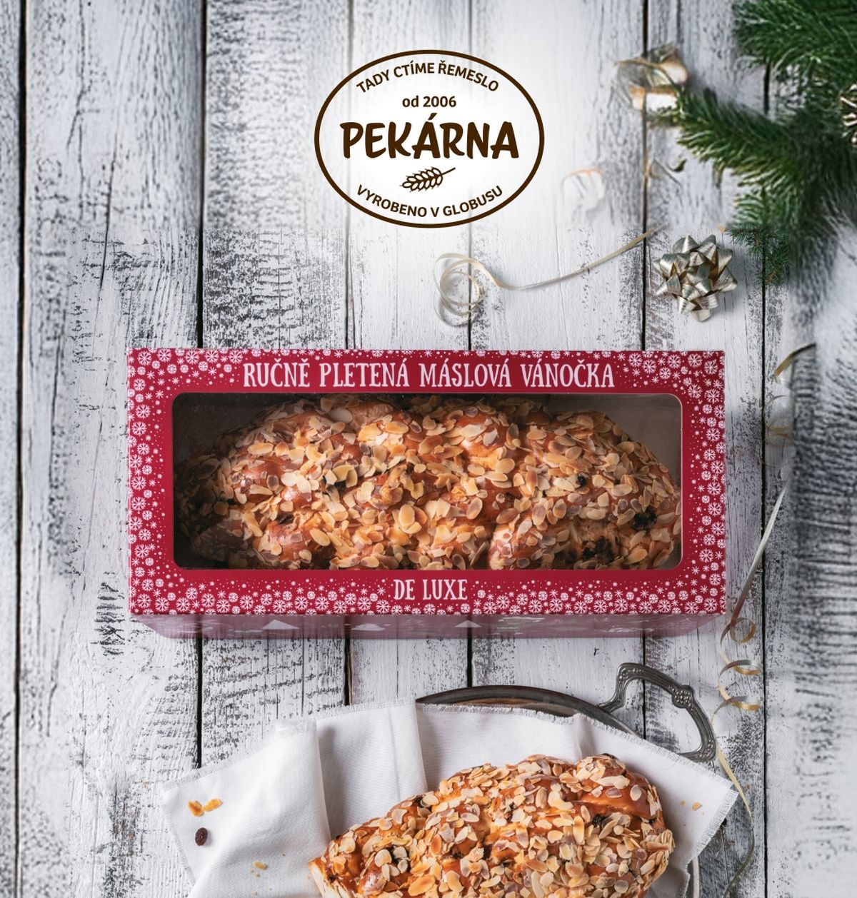 Máslová vánočka de luxe v červené papírové krabici, kus rozkrojené vánočky na bílé látce a tácu, zelené jehličí s ozdobami, vše na dřevěném pozadí