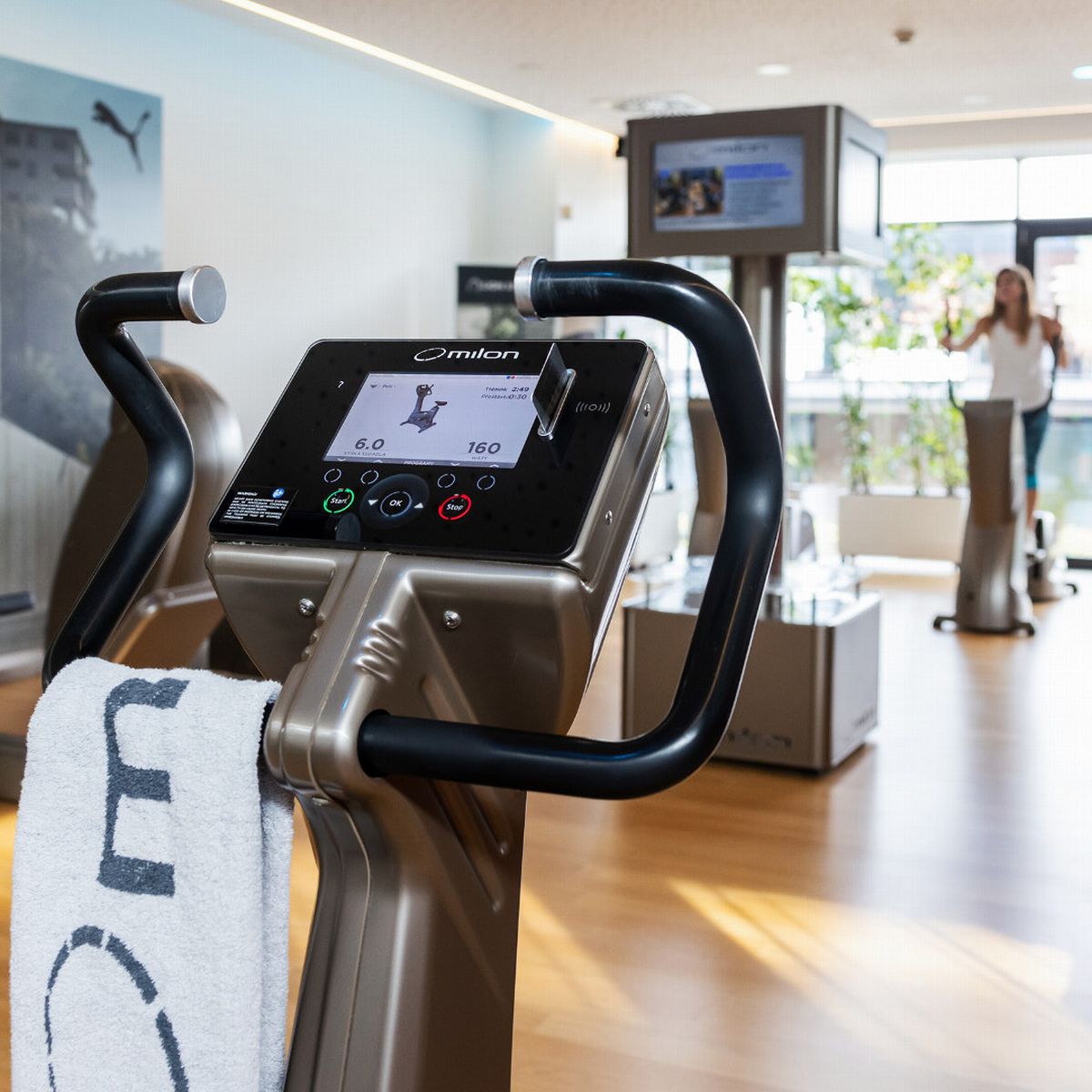 Moderní posilovna s elegantními posilovacími stroji, velkými okny pro přirozené světlo a ručníkem přehozeným přes řídítka stacionárního kola.