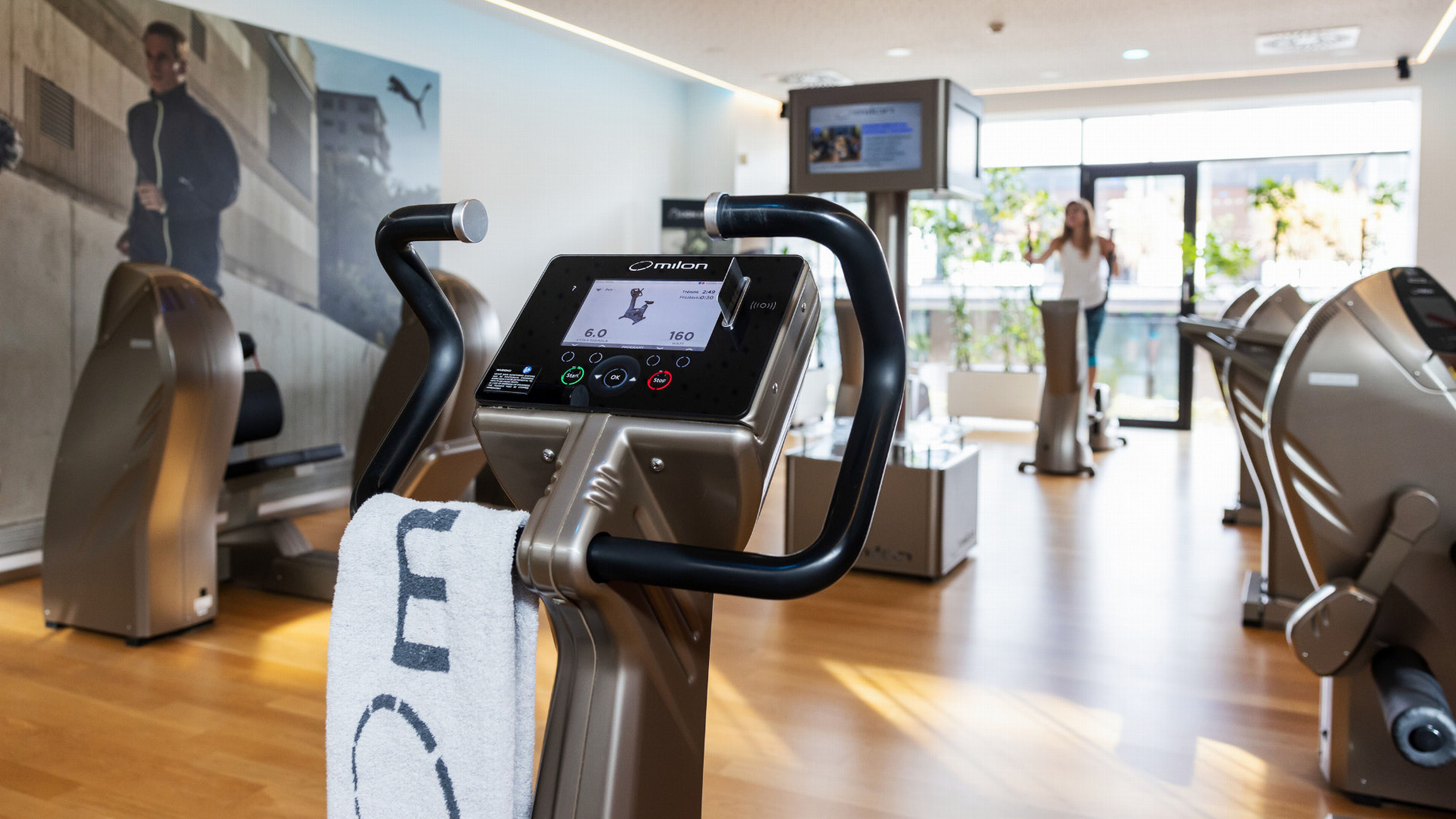 Moderní posilovna s elegantními posilovacími stroji, velkými okny pro přirozené světlo a ručníkem přehozeným přes řídítka stacionárního kola.