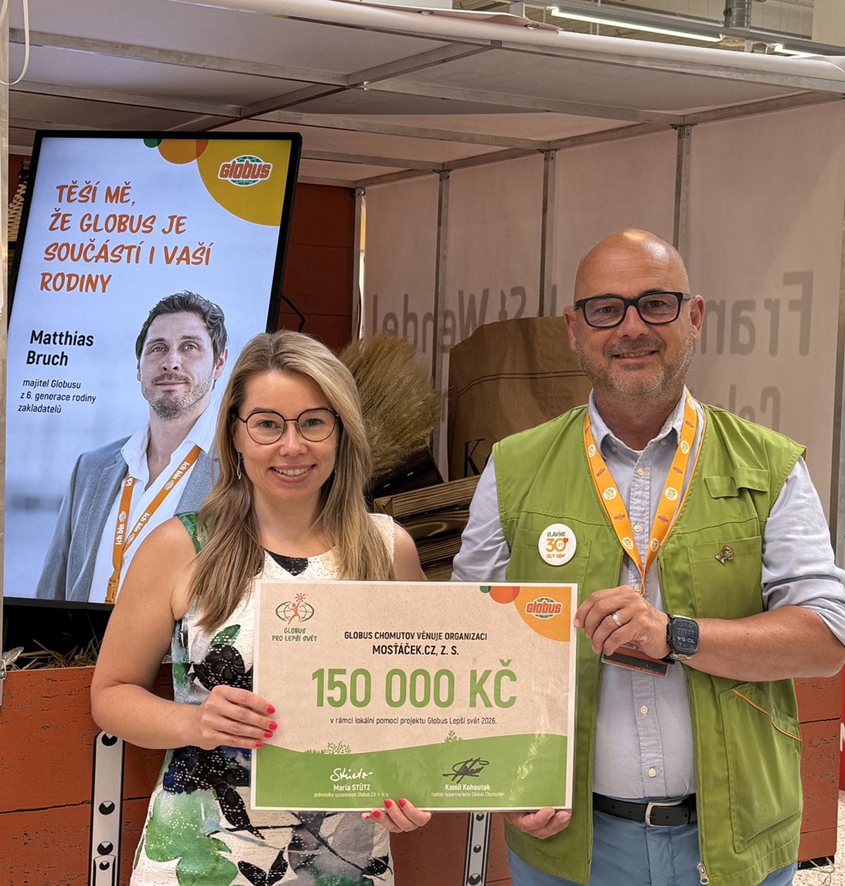 Dvě osoby drží v obchodě velký šek na 150 000 Kč, v pozadí je plakát a potraviny.