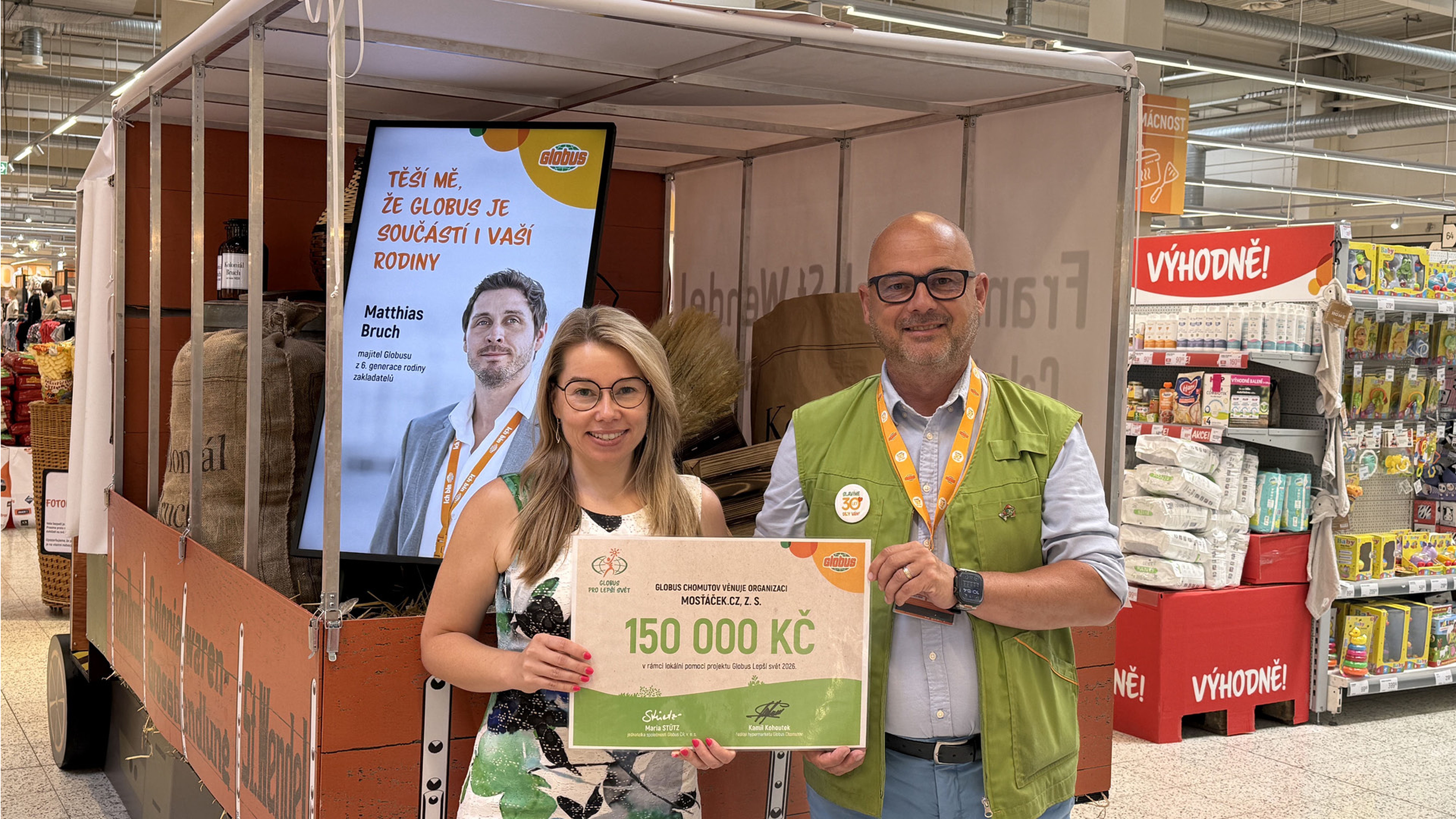 Dvě osoby drží v obchodě velký šek na 150 000 Kč, v pozadí je plakát a potraviny.