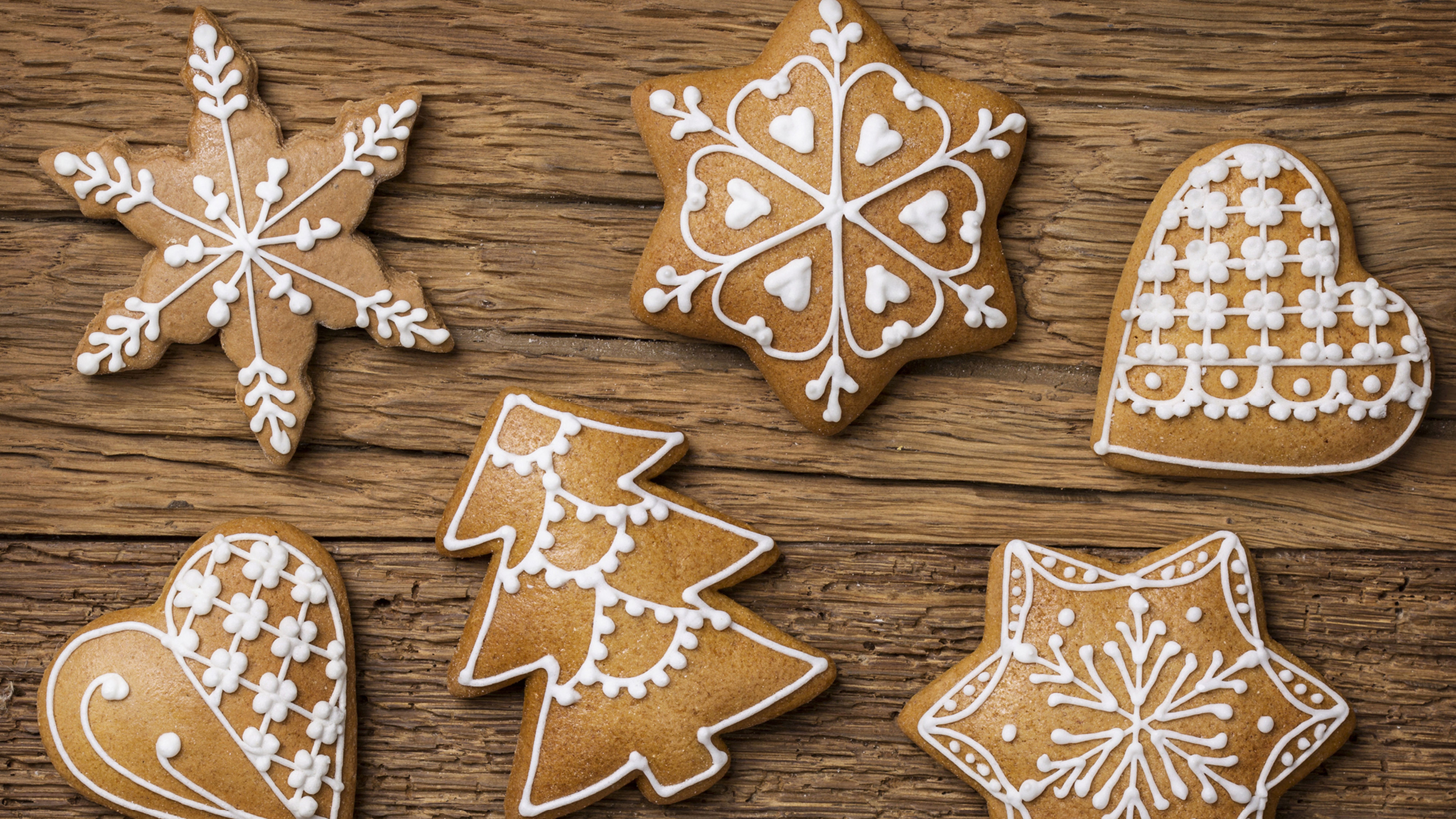 Na hnědém dřevěném podkladu jsou perníčky ve tvaru vločky, hvězdy, srdce a stromečku ozdobené bílou polevou.